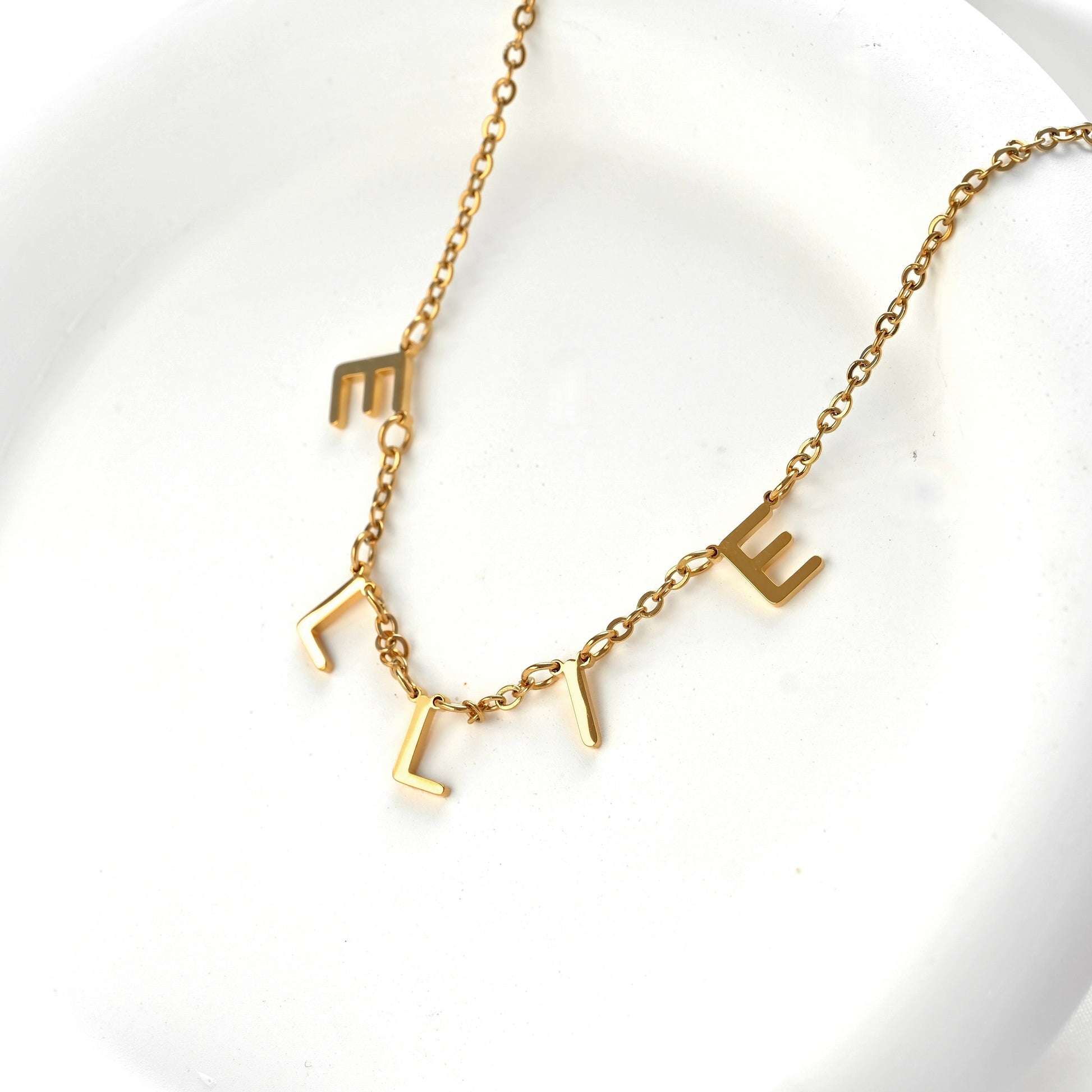 A gold initial necklace spelling 'ELLIE' with letter-shaped charms on a delicate chain, displayed on a white textured surface.