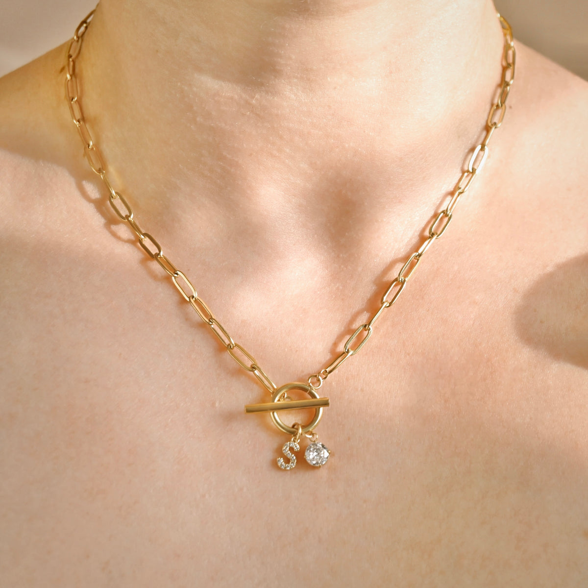 A close-up of a gold chain necklace with a toggle clasp and two small dangling charms, worn on a model's neck.