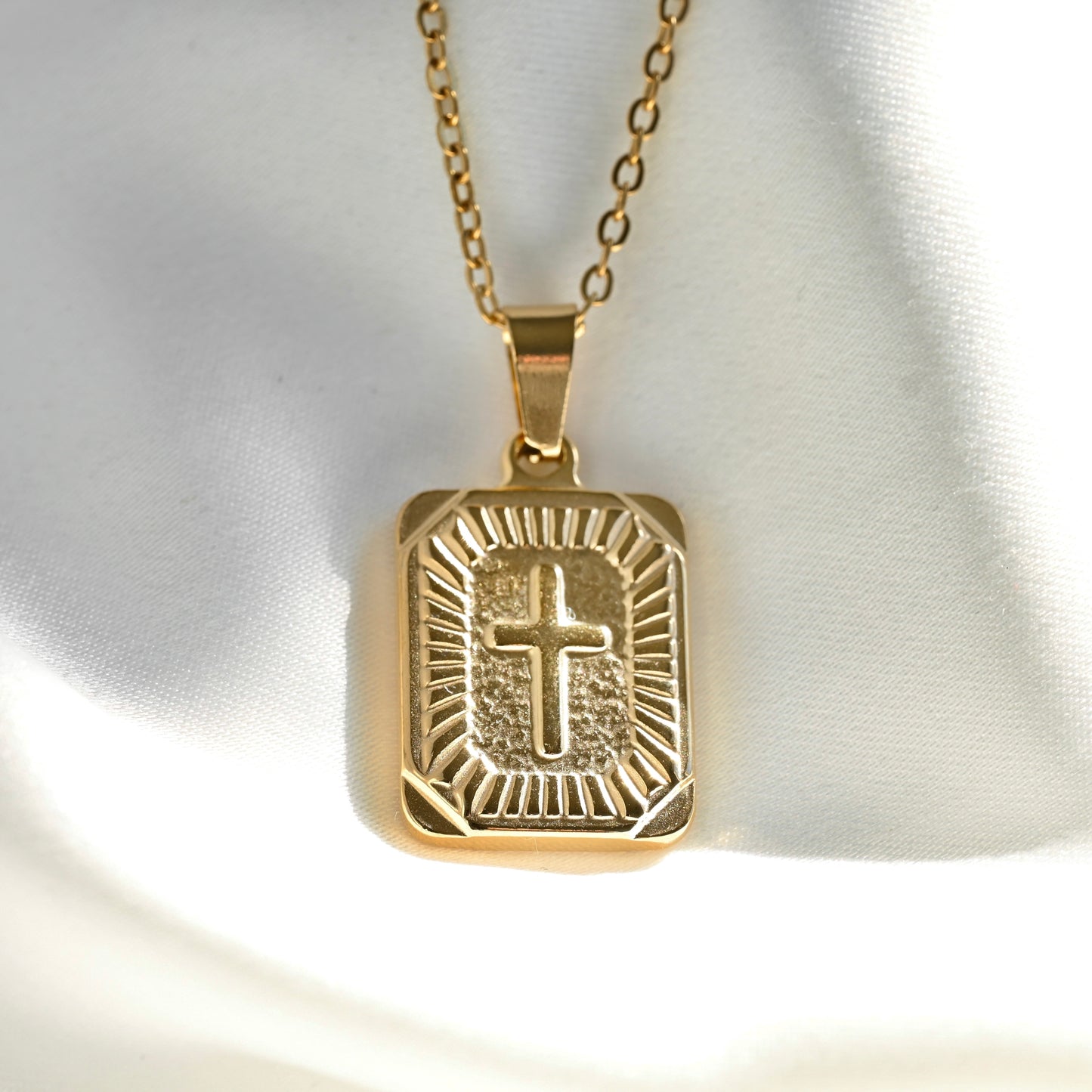A gold rectangular pendant necklace with a raised cross and sunburst design, resting on a white satin fabric background.