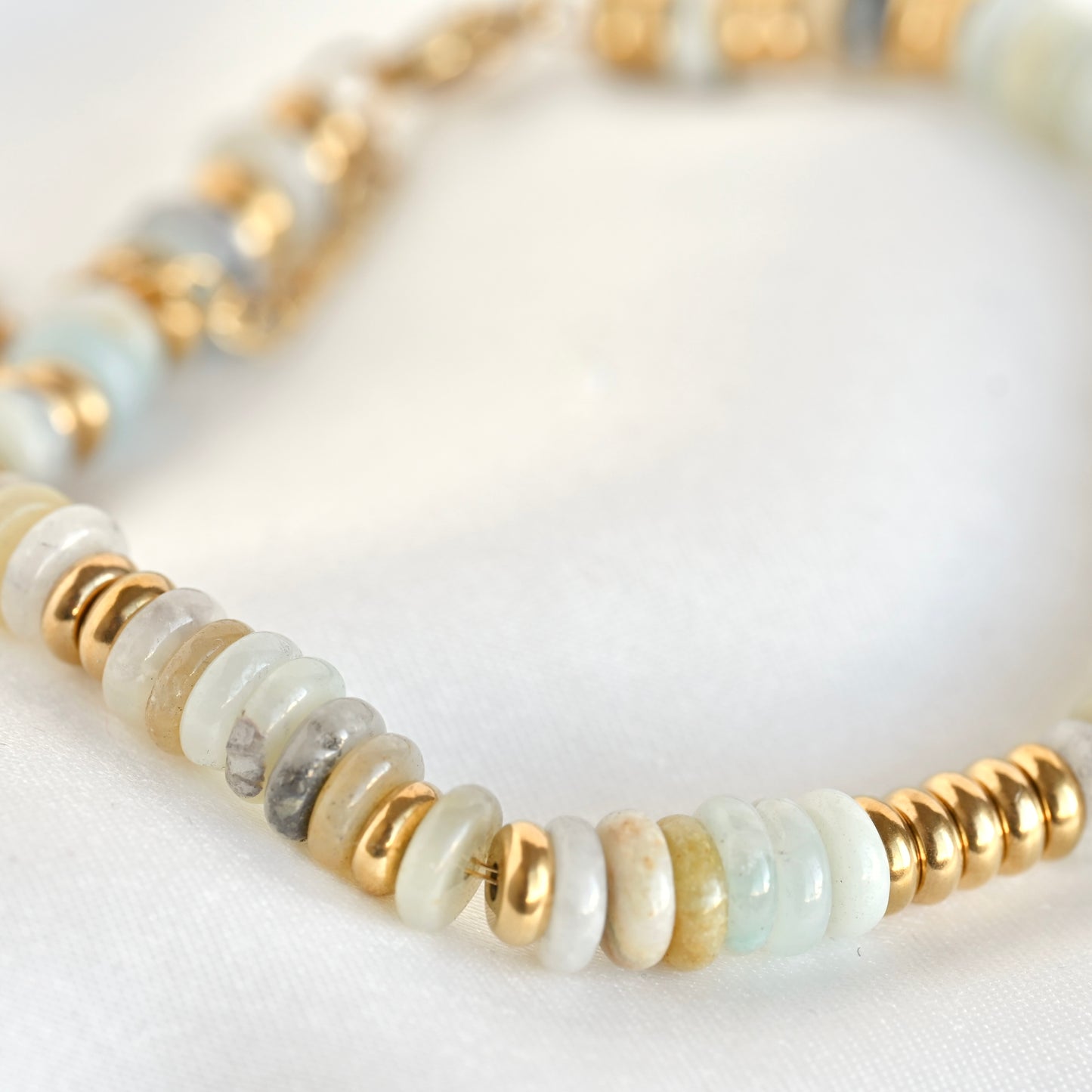A close-up of a beaded bracelet featuring round white and pale blue stone beads, light tan beads, and shiny gold spacer beads on a soft white fabric background.