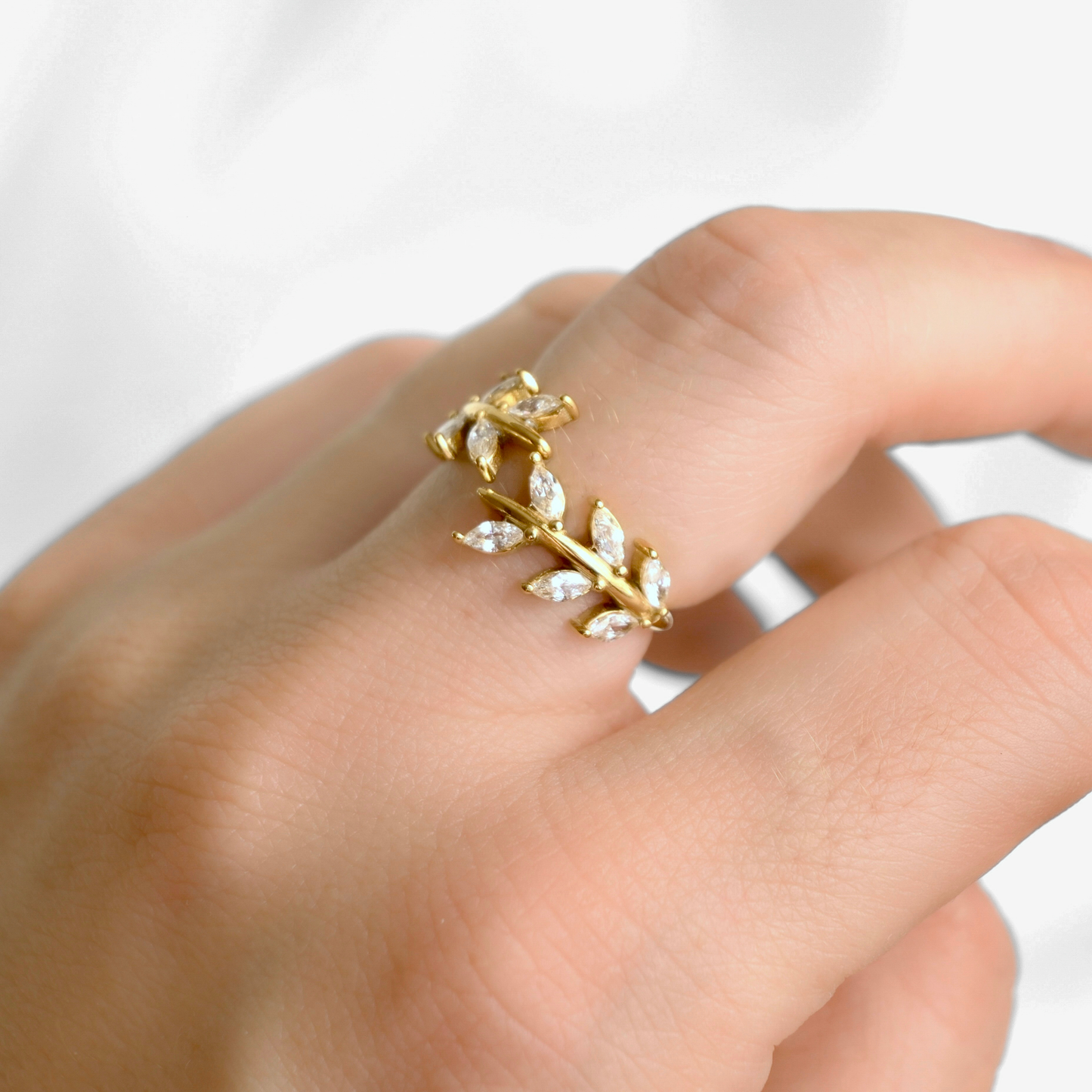 A gold leaf-shaped bypass ring with marquise-cut clear stones, shown worn on a person's finger against a soft white background.