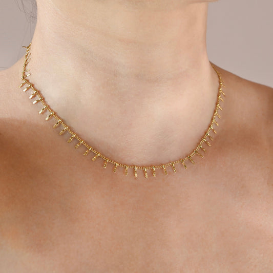 A delicate gold necklace with small dangling bead-like charms, shown worn on a model's neck against a soft beige background.