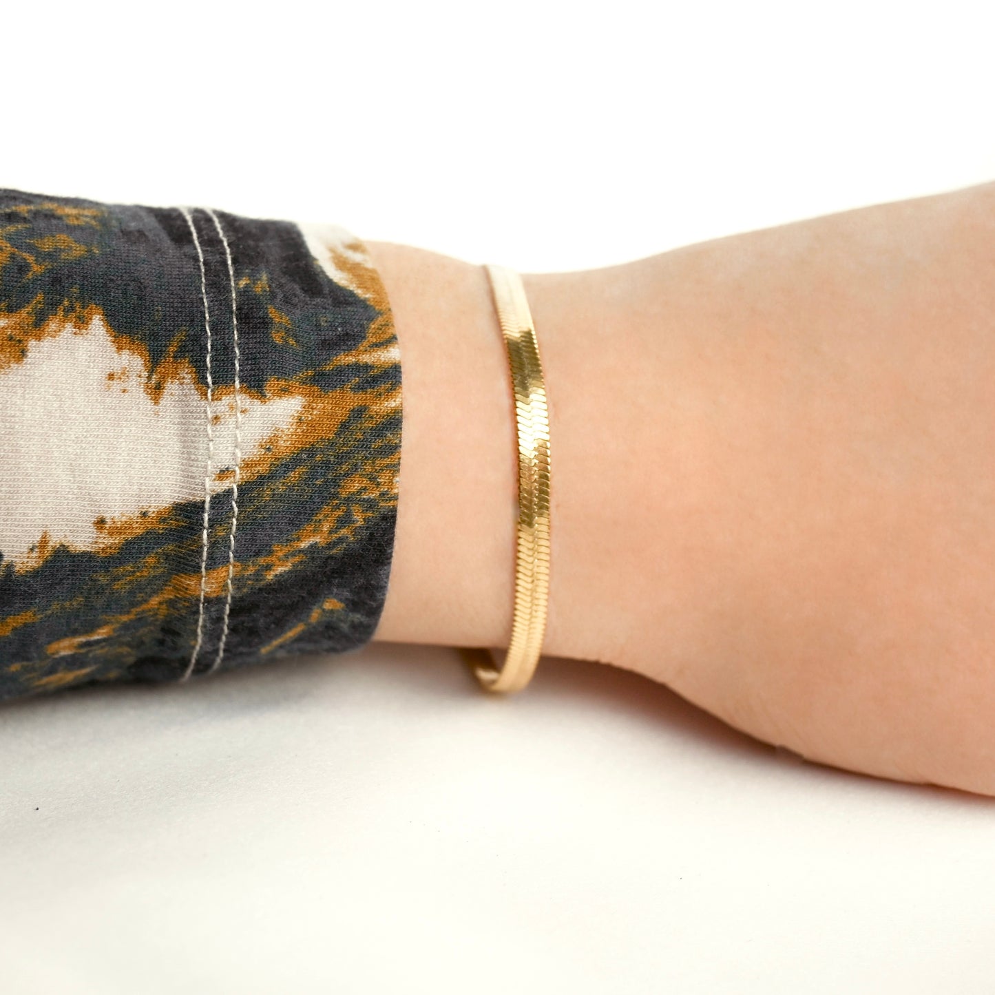 Gold 4mm herringbone bracelet on a wrist with a patterned sleeve against a white background