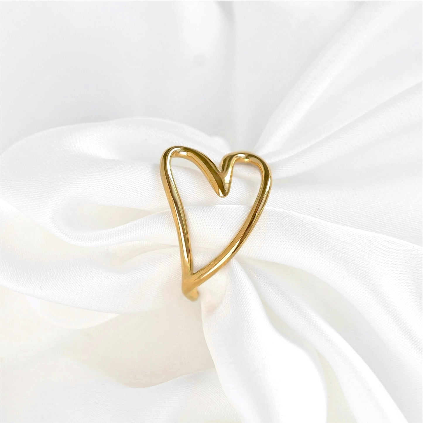 A gold heart-shaped ring with an open heart design, displayed on a white satin fabric.