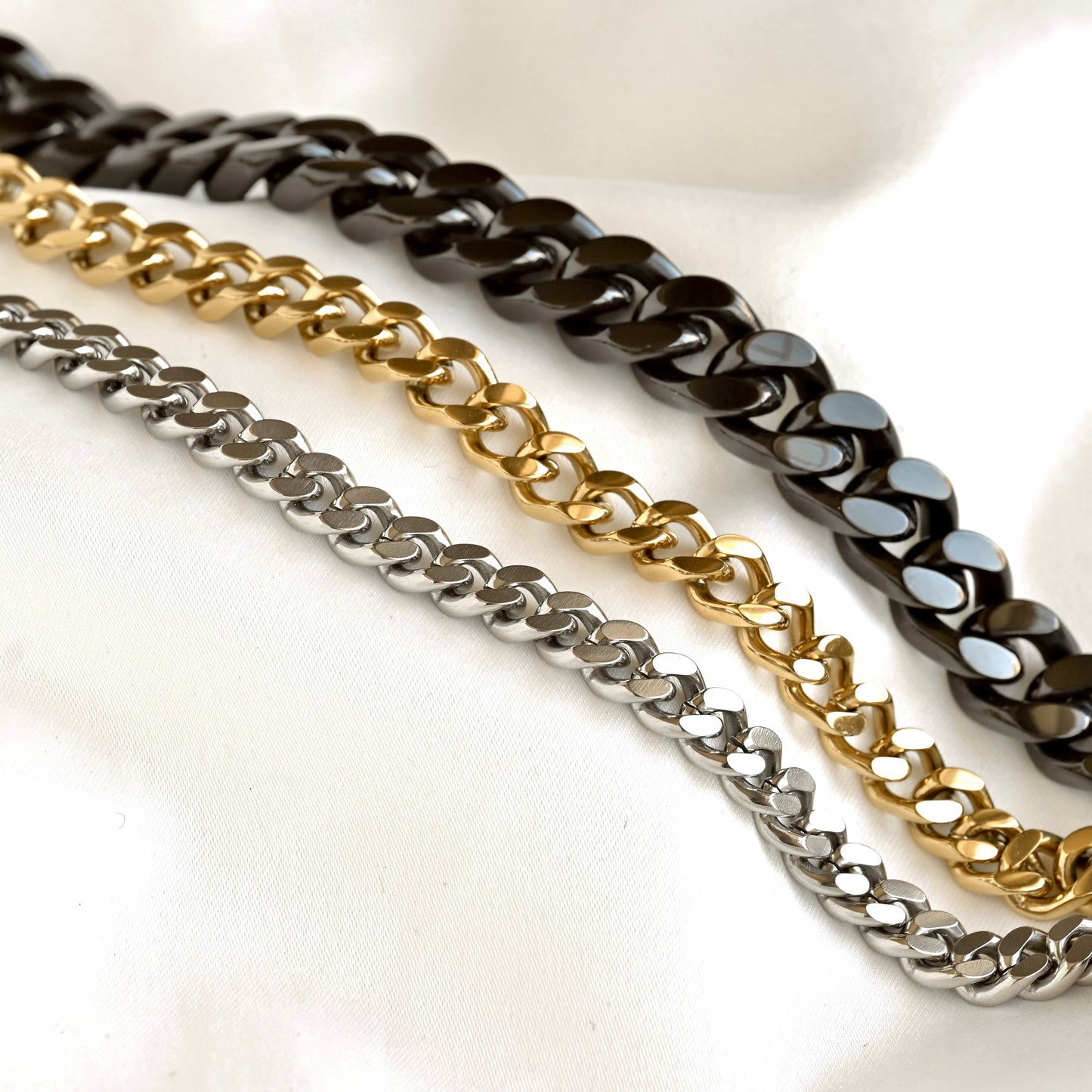 Three metallic cuban chain straps, including a black chain, a gold chain, and a silver chain, arranged diagonally on a white fabric background.