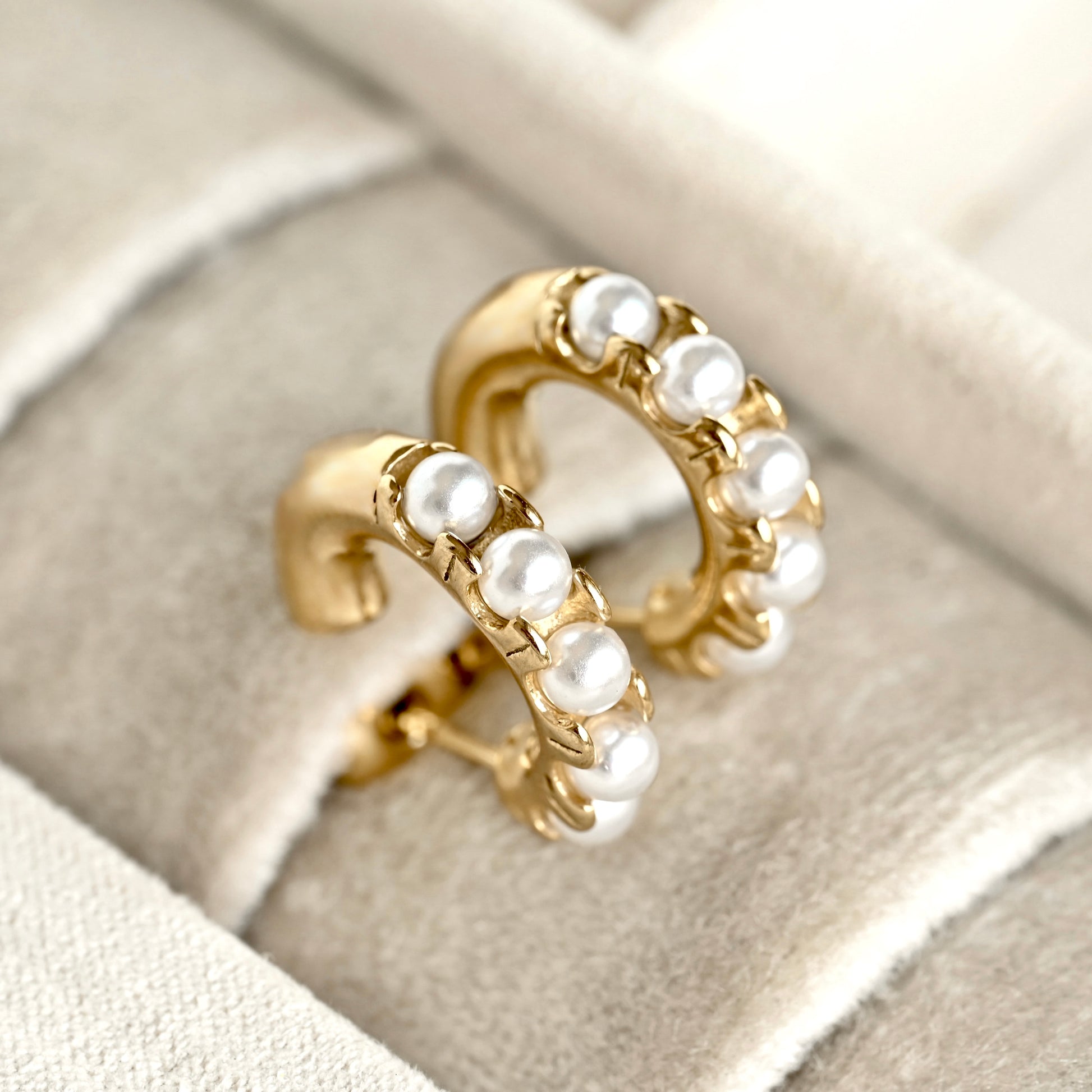 A pair of gold hoop earrings adorned with white faux pearls, displayed on a beige velvet jewelry box.
