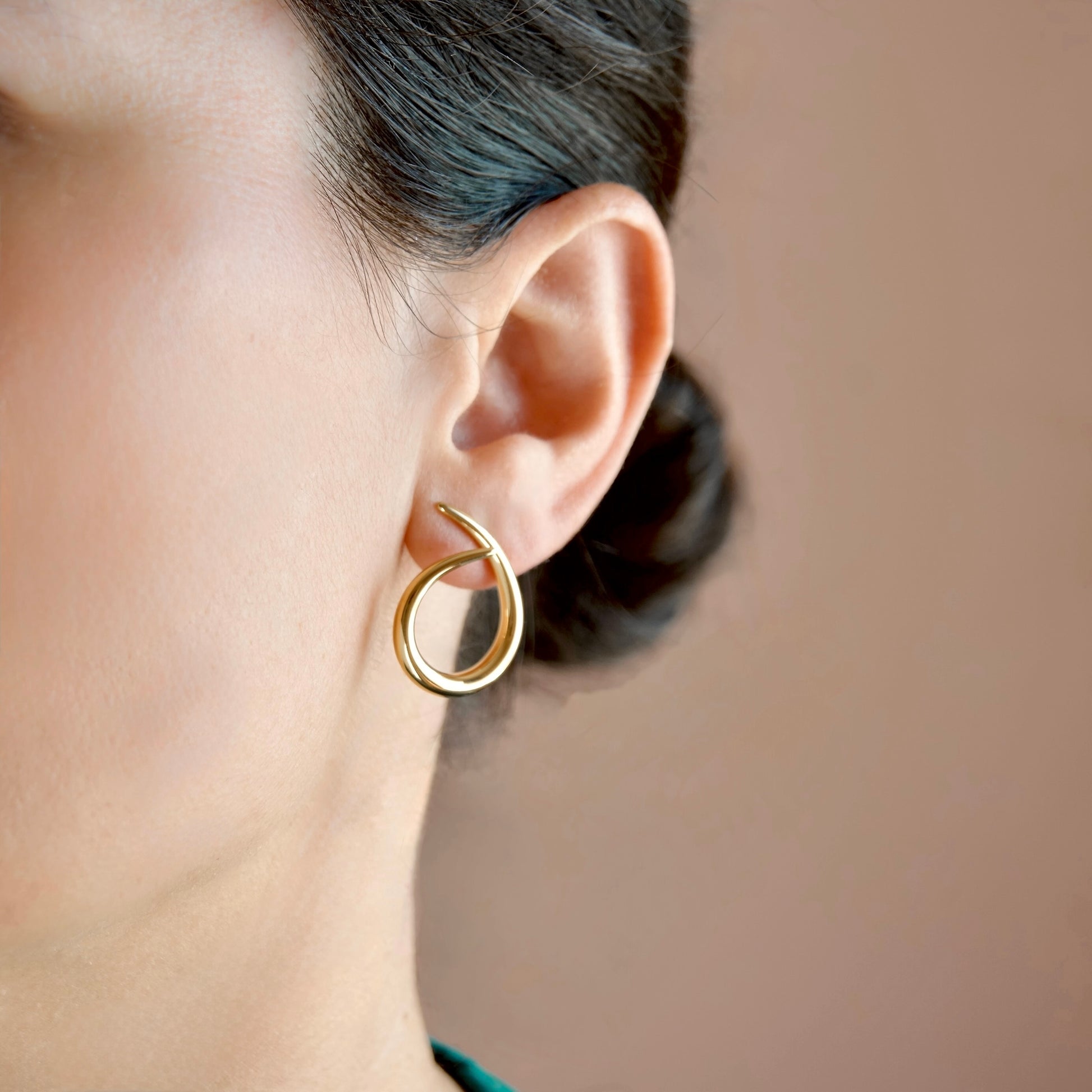 A close-up of a person's ear wearing a polished gold crescent-shaped hoop earring against a soft warm beige background.