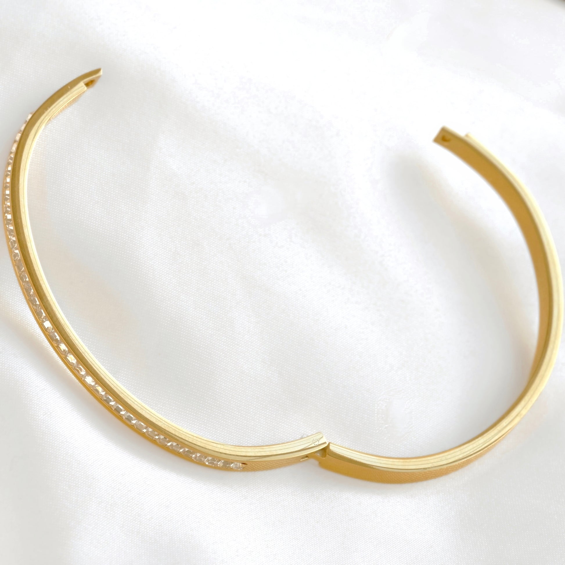 A gold-toned open hinged bangle with a row of small clear stones along one side, displayed on a soft white fabric background.