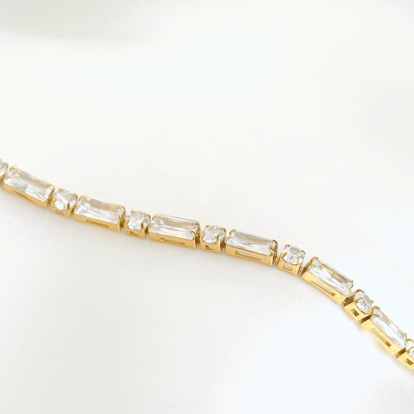 A close-up of a yellow gold tennis bracelet featuring baguette and round clear crystals, set against a clean white background.