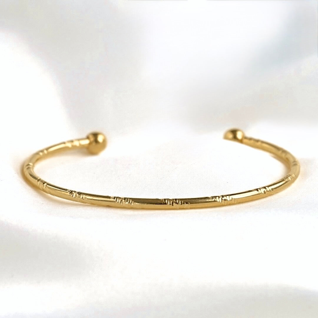 A thin gold open cuff bracelet with a textured, segmented look and ball ends, displayed on a soft white fabric background.