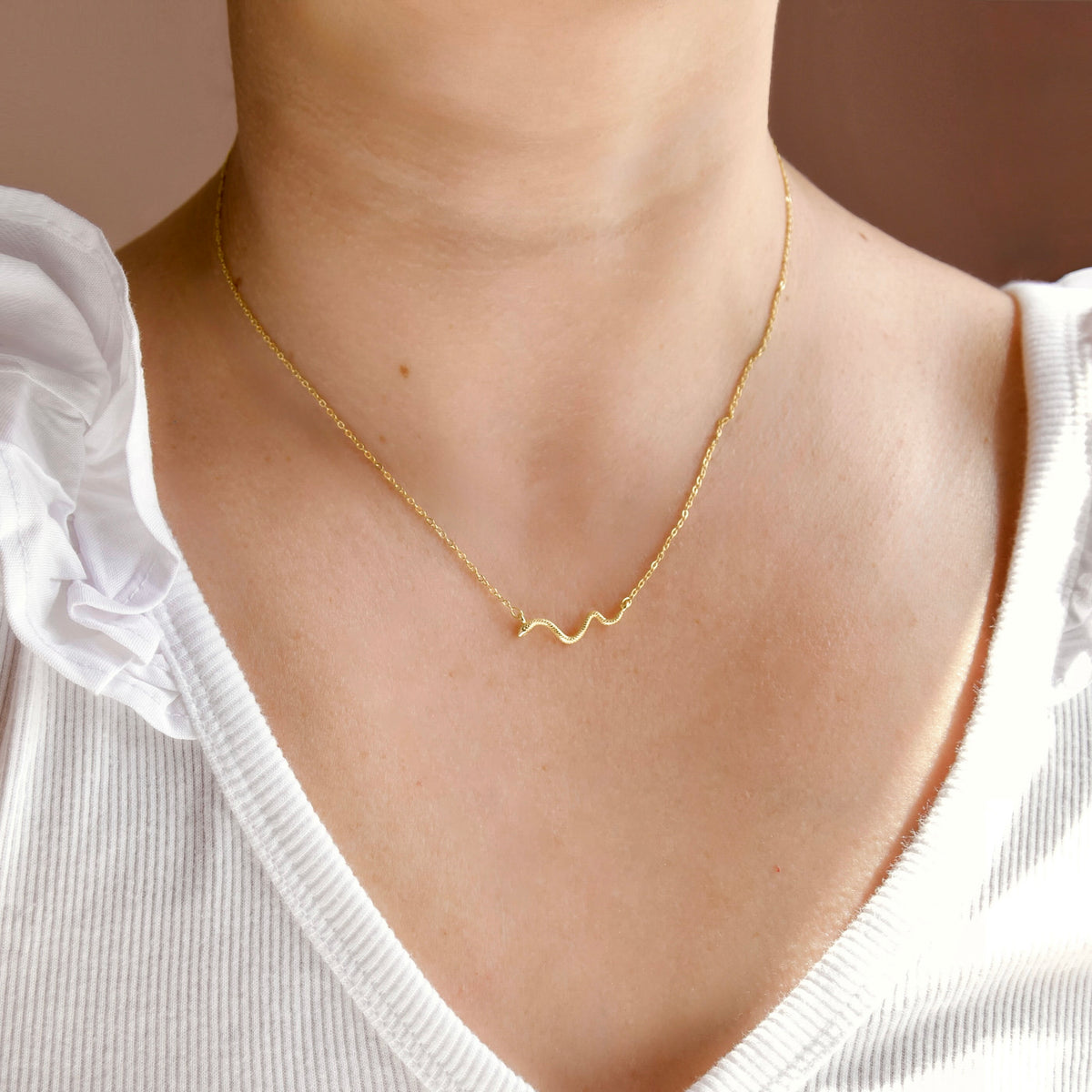 A close-up of a person's neck and upper chest wearing a delicate gold chain necklace with a small wavy pendant, paired with a white ribbed top.