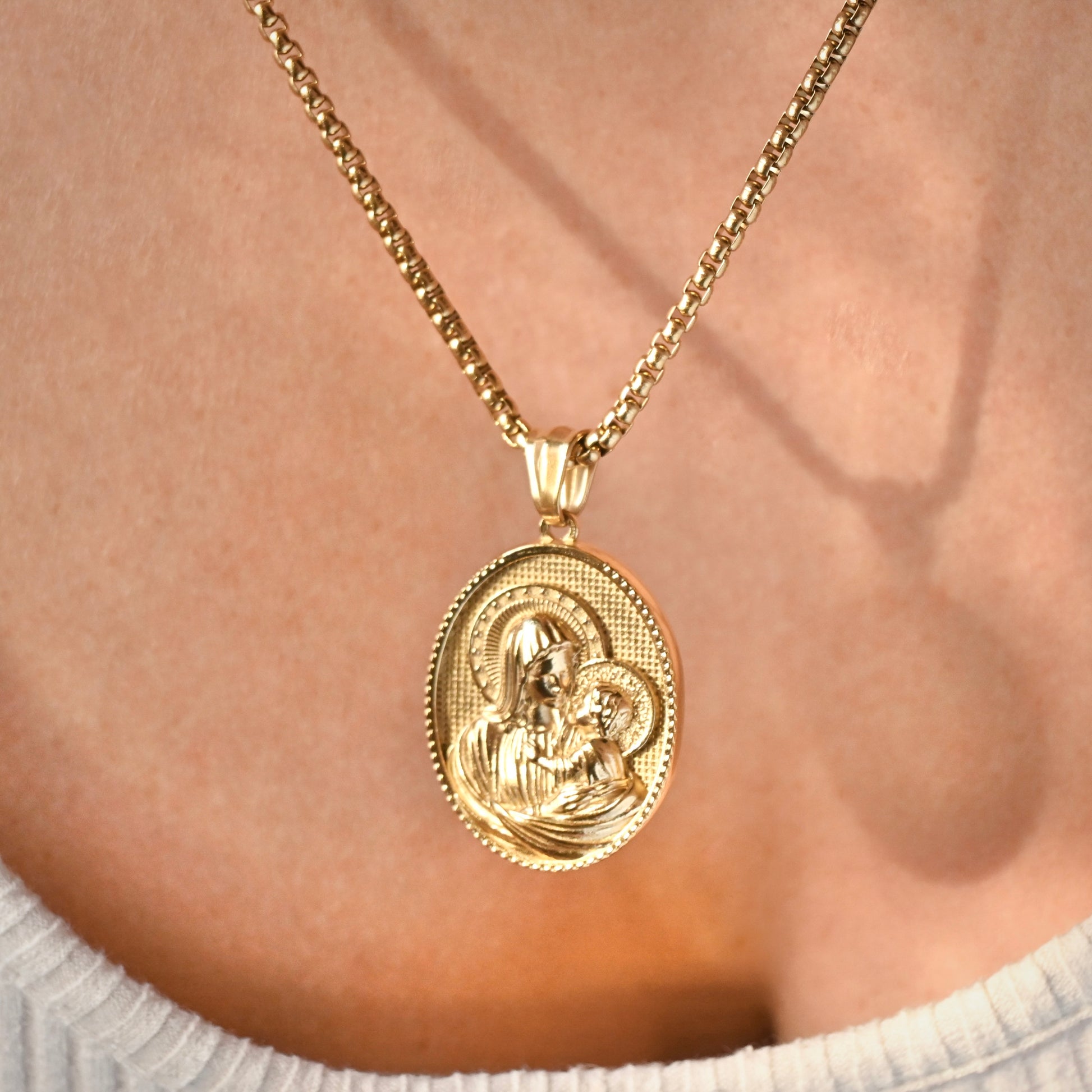 A gold oval pendant necklace featuring an embossed religious icon, worn on a gold chain over a person's chest.
