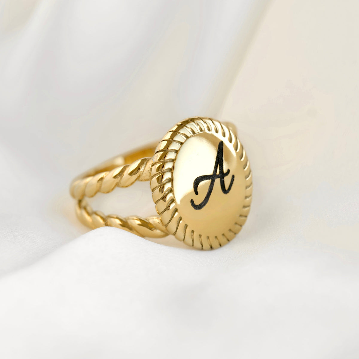A gold signet ring with a twisted rope band and an oval face engraved with a black capital letter 'A', resting on draped white fabric.