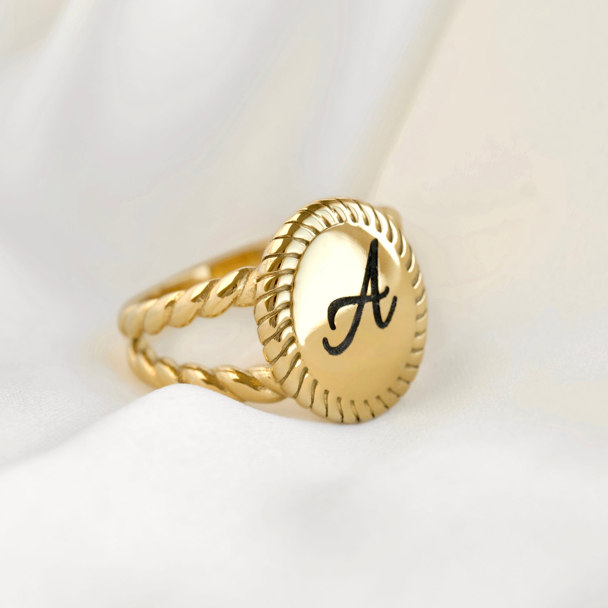 A gold signet ring with a twisted rope band and an oval face engraved with a black capital letter 'A', resting on draped white fabric.