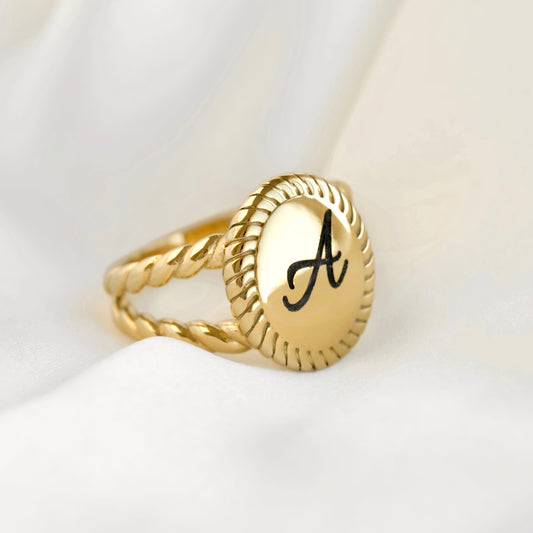 A gold signet ring with a twisted rope band and an oval face engraved with a black capital letter 'A', resting on draped white fabric.