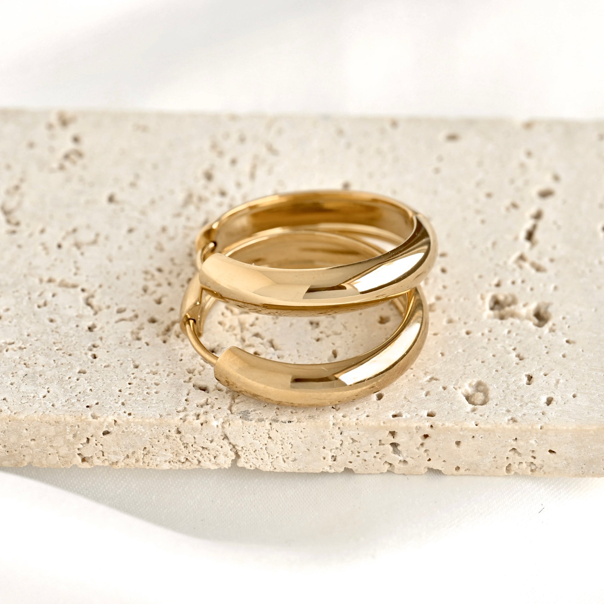 Two polished gold huggie rings stacked together, displayed on a textured beige stone slab against a clean white background.