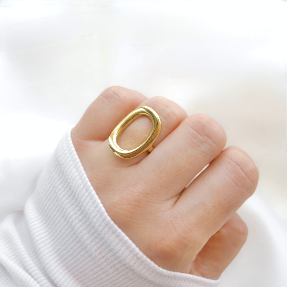 A close-up of a hand wearing a thick, polished gold ring with a rounded, open oval shape, styled with a white ribbed knit sleeve.