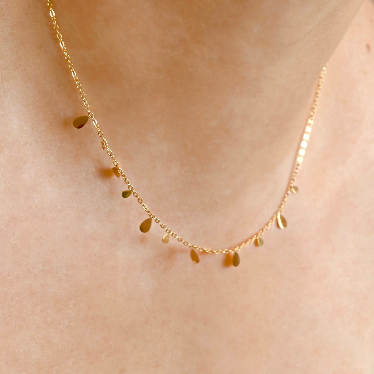 A delicate gold chain necklace with small teardrop-shaped charms, shown worn on a person's neck against a warm skin tone background.