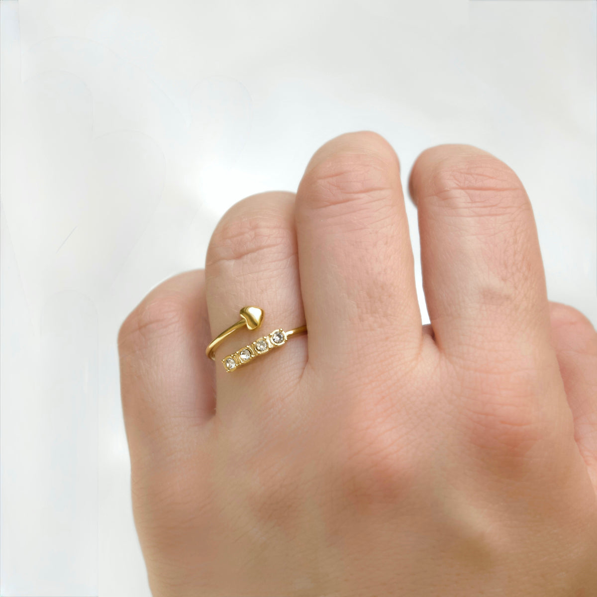 A close-up of a hand wearing a delicate gold ring with a small heart topper and a row of clear stones on a white background.