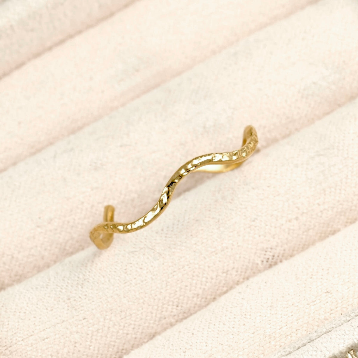 A textured gold wavy ring with a small heart-shaped accent on the band, displayed on a light beige textured fabric background.