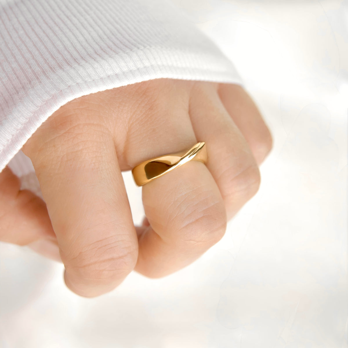 A close-up of a hand wearing a polished gold ring with a unique twisted, looped design, styled with a white ribbed knit sleeve.