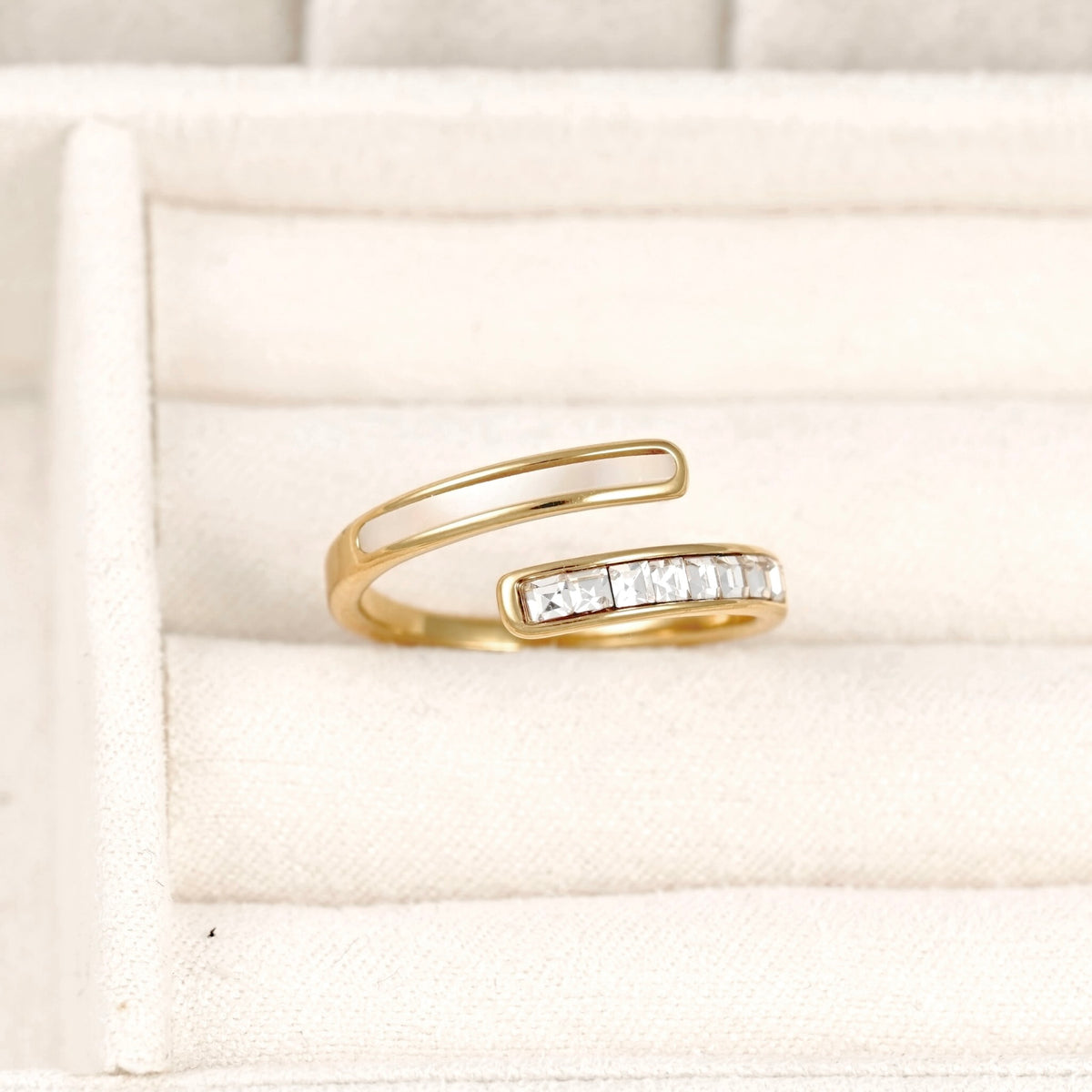 A gold open-band ring with a row of clear rectangular stones on one side, displayed on a cream-colored jewelry box cushion.