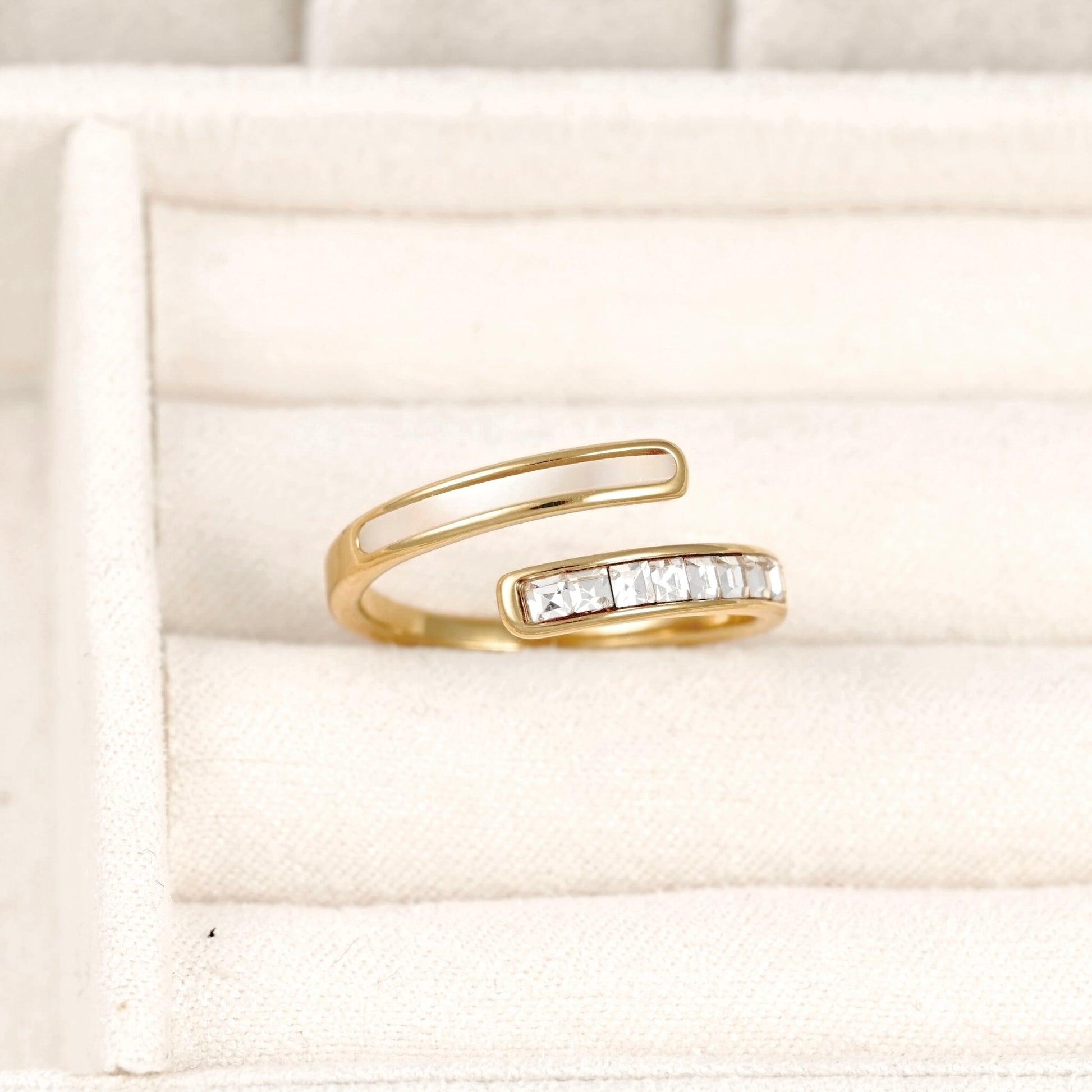 A gold open-band ring with a row of clear rectangular stones on one side, displayed on a cream-colored jewelry box cushion.