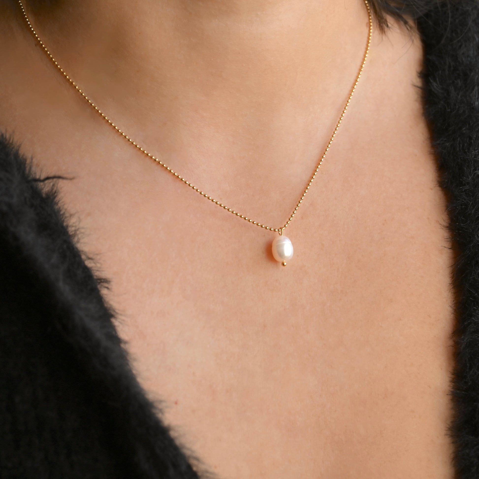 A delicate gold chain necklace with a single small white pearl pendant, worn on a model's neck over a black fuzzy top.