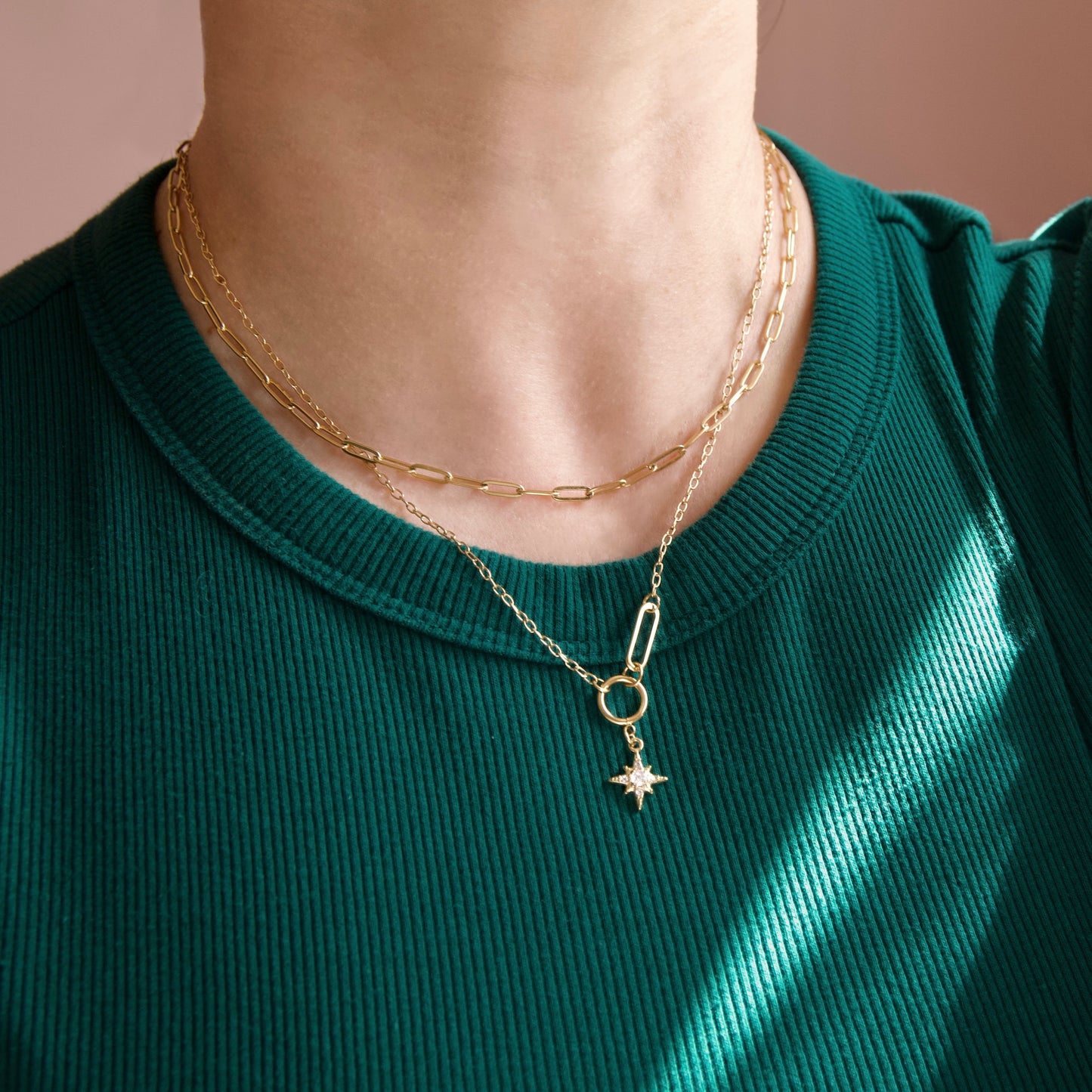 A close-up of a person's neck and chest wearing a layered gold necklace set over a dark green ribbed top. The necklace features a paperclip charm with a star and a small circular ring.