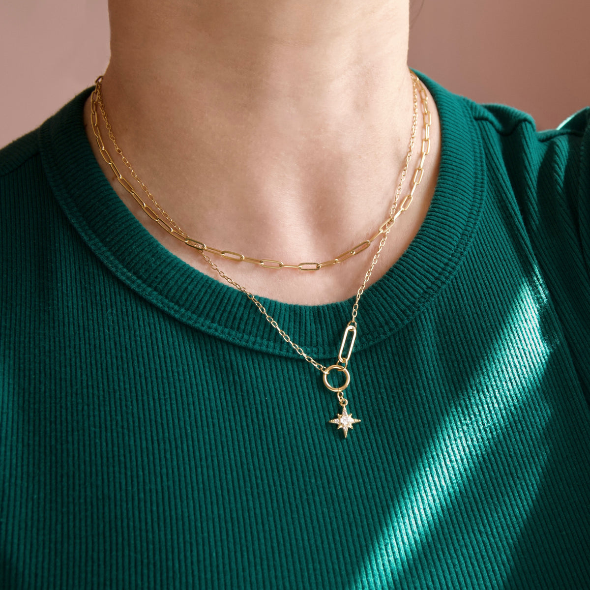 A close-up of a person's neck and chest wearing a layered gold necklace set over a dark green ribbed top. The necklace features a paperclip charm with a star and a small circular ring.