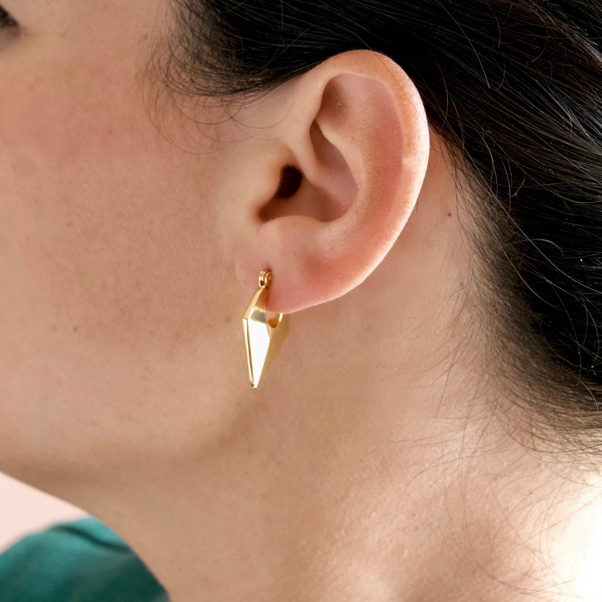 A close-up of a person's ear wearing a small geometric gold dangle earring.