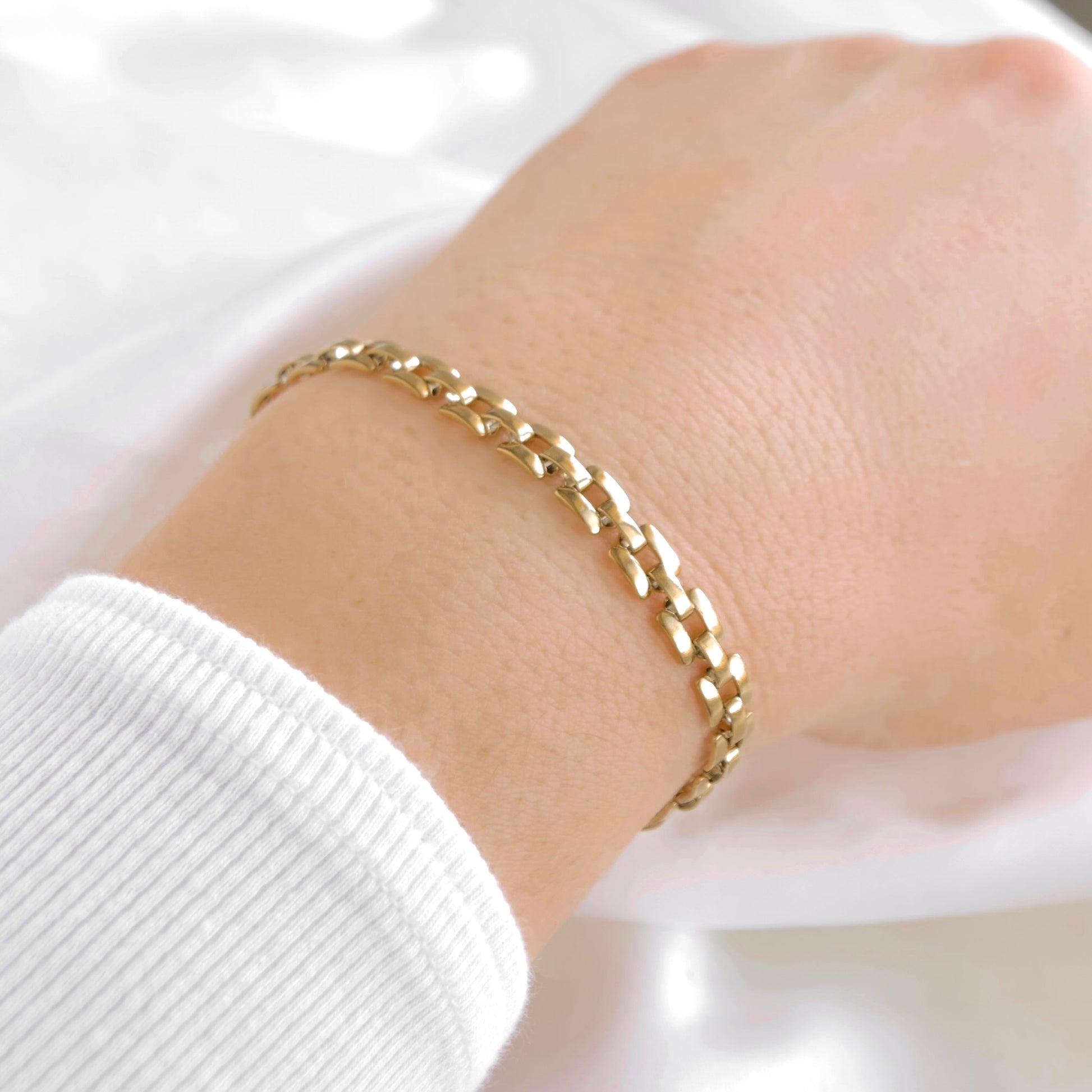 A close-up of a person's wrist wearing a delicate gold curb chain bracelet, paired with a white ribbed long-sleeve top against a soft white fabric background.