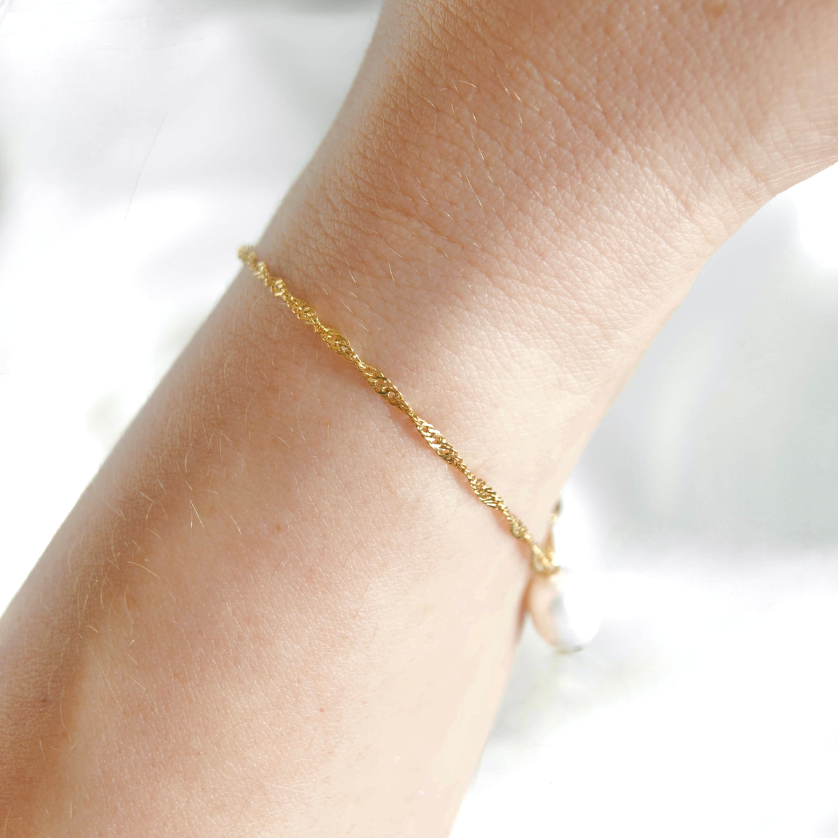 A delicate gold chain bracelet with a small white pearl charm, shown worn on a person's wrist against a soft white background.