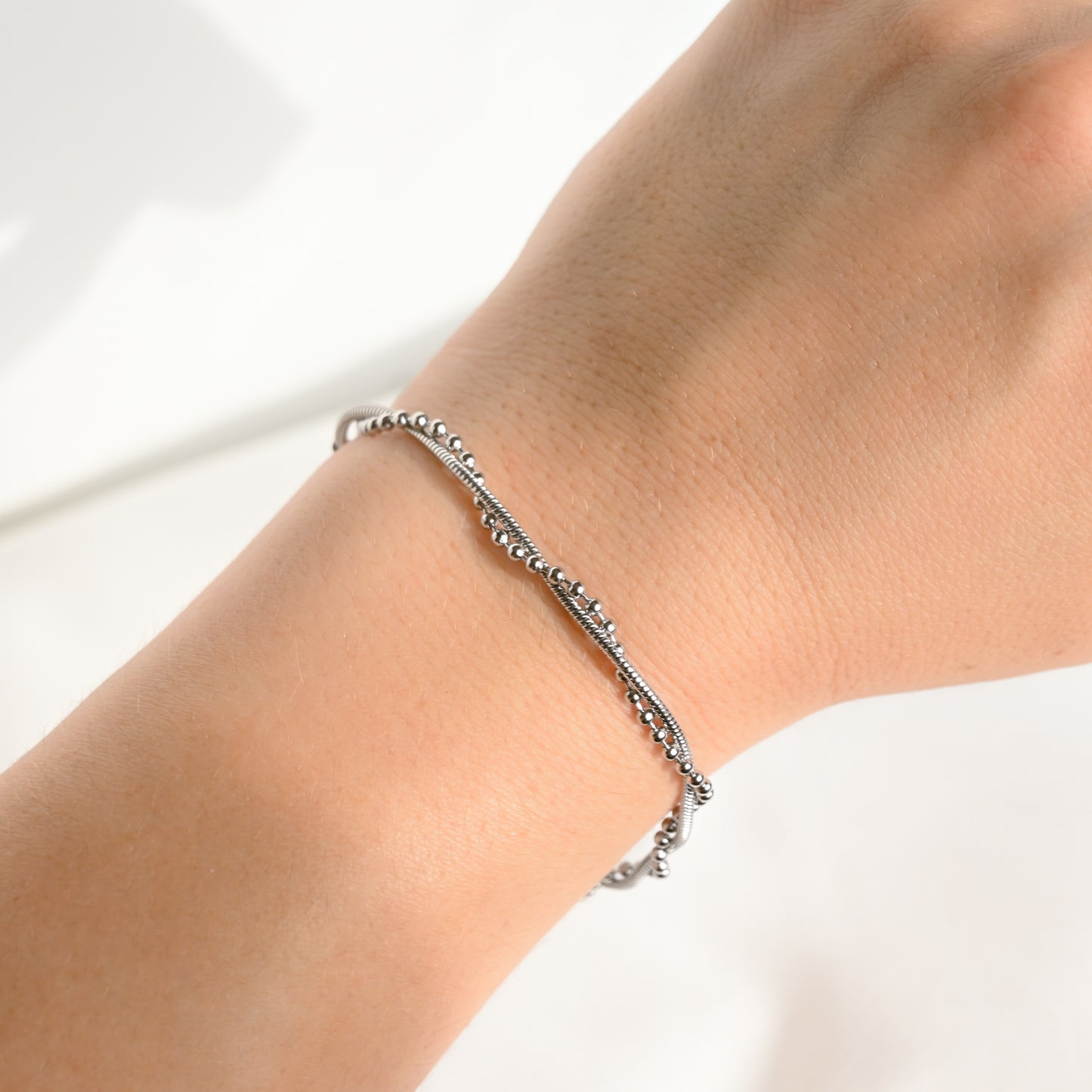 A close-up of a delicate silver beaded bracelet worn on a person's wrist against a bright white background.