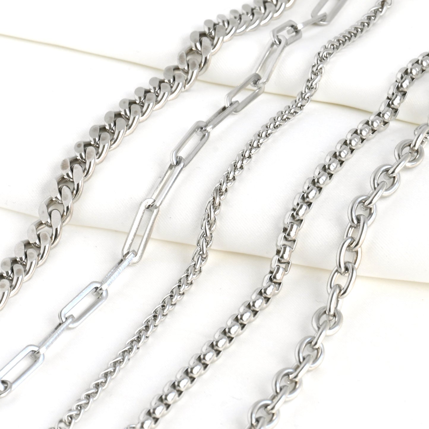 Collection of silver chains on a light background. From left to right: Cuban, paperclip, wheat, rounded box and rolo