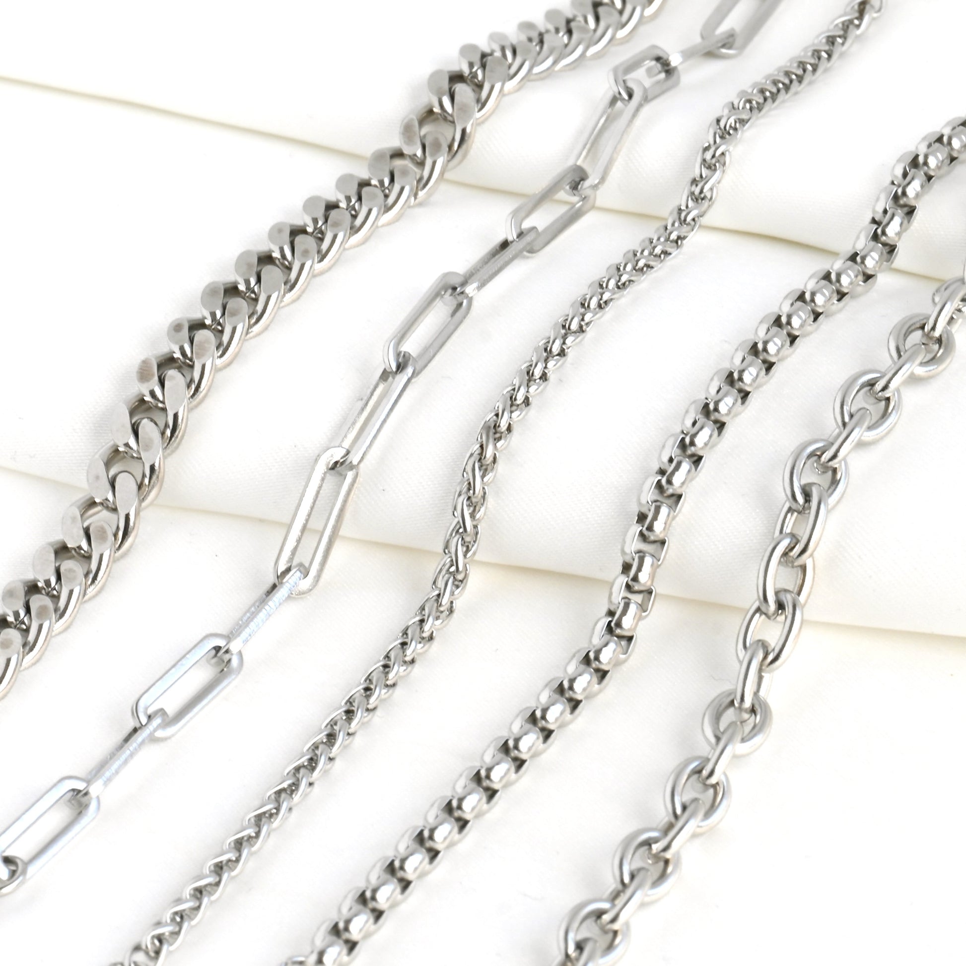 Collection of silver chains on a light background. From left to right: Cuban, paperclip, wheat, rounded box and rolo