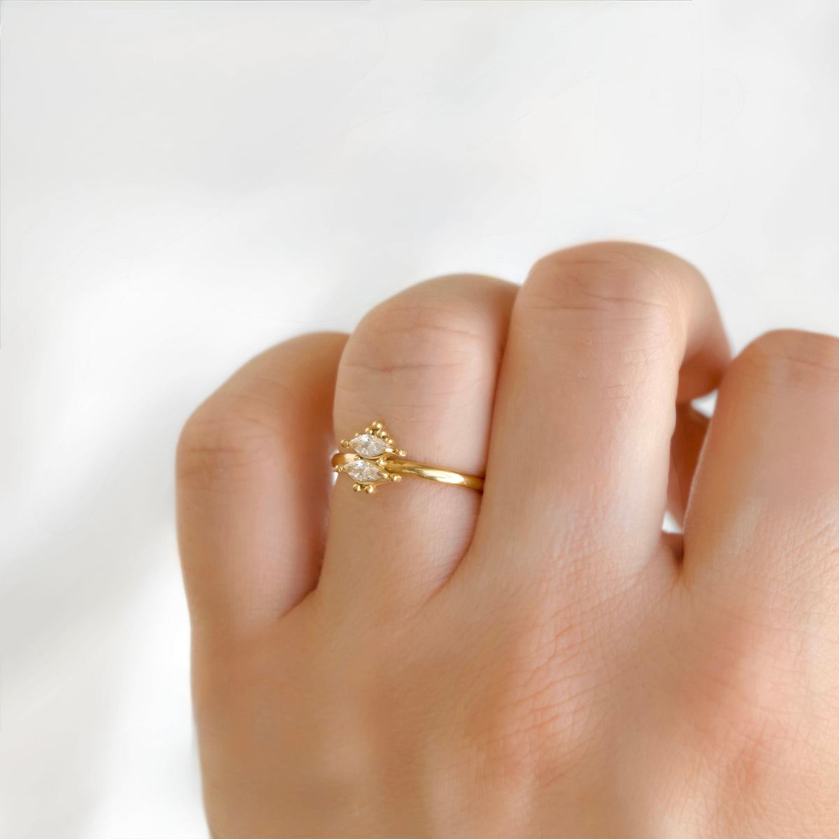 A gold ring with a small clear gemstone set in a decorative cluster, worn on a person's ring finger against a soft white background.