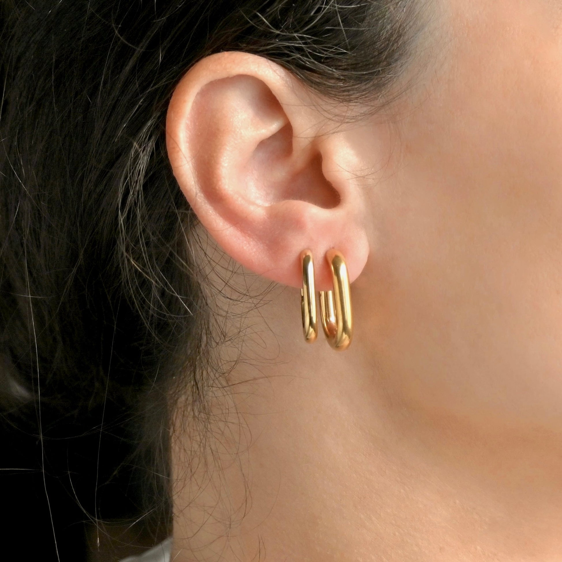 Pair of gold  U hoops, one thick with C-shape, other thin and hinged worn by a person with dark hair.