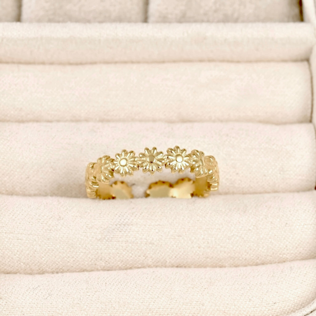A gold ring featuring a continuous row of small daisy-like flowers, displayed on a cream-colored textured ring box.