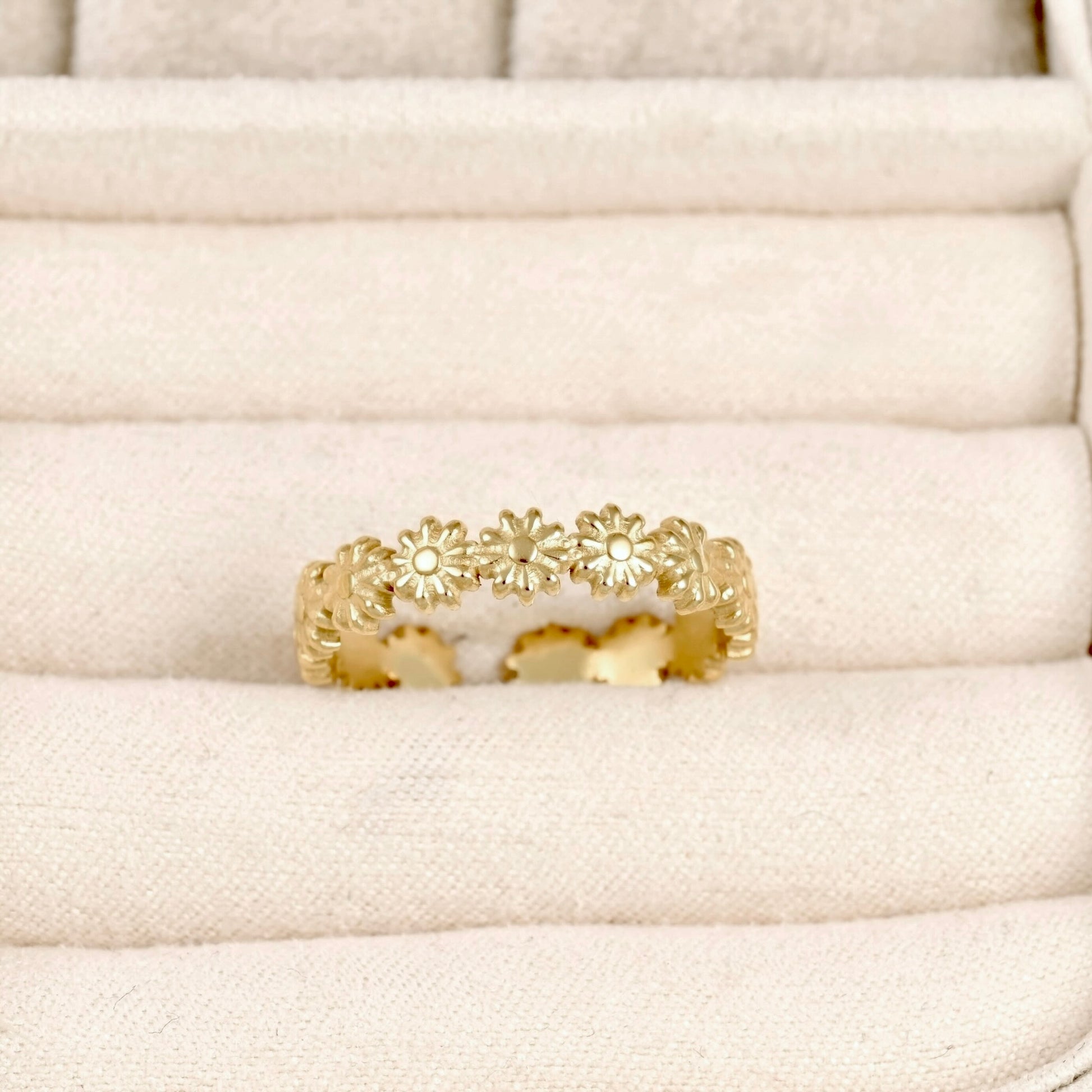 A gold ring featuring a continuous row of small daisy-like flowers, displayed on a cream-colored textured ring box.