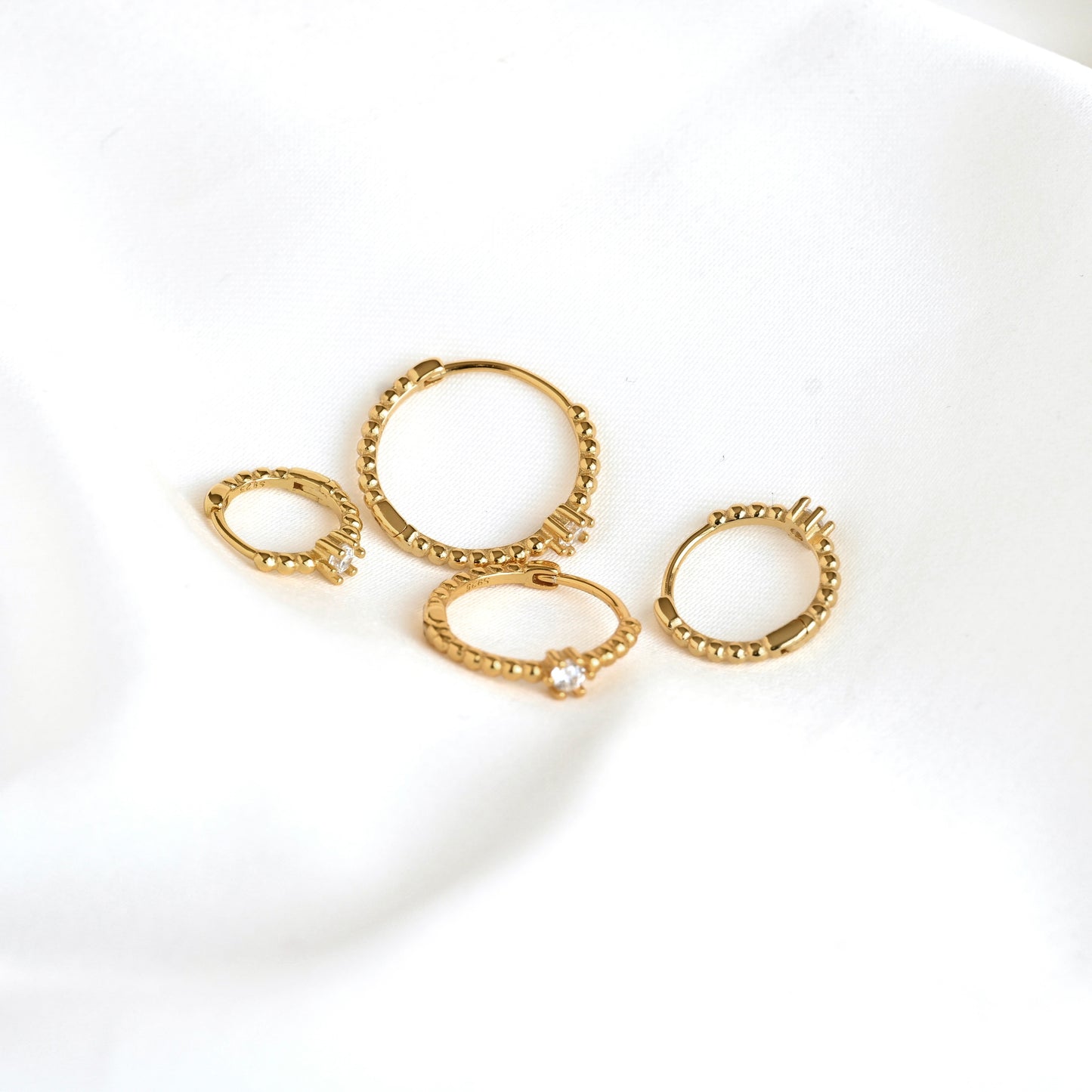 Four gold-toned hoop earrings with textured beaded edges and small clear stones, arranged on a soft white fabric background.