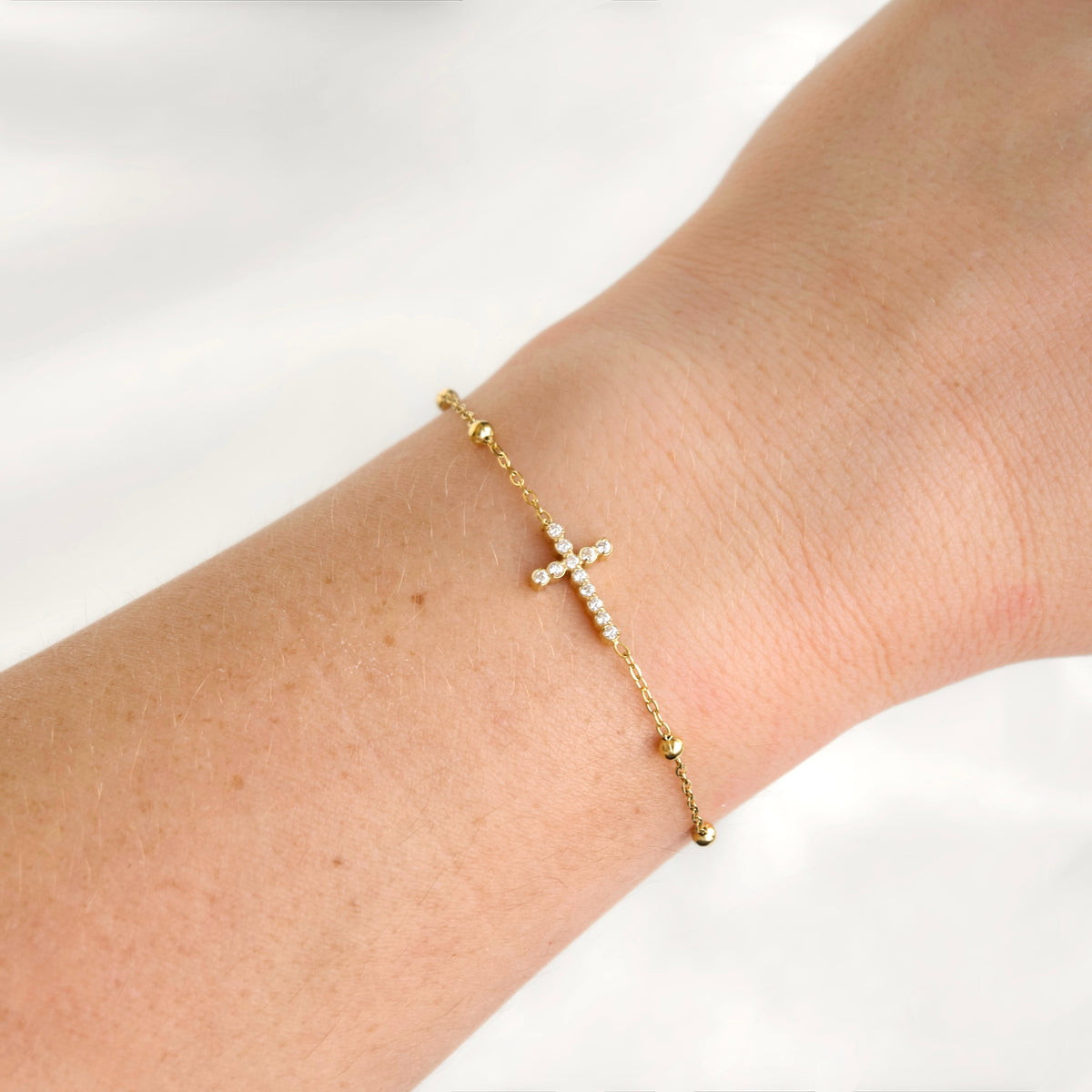 A delicate gold chain bracelet with a small cross charm set with clear stones, shown worn on a person's wrist against a soft white background.