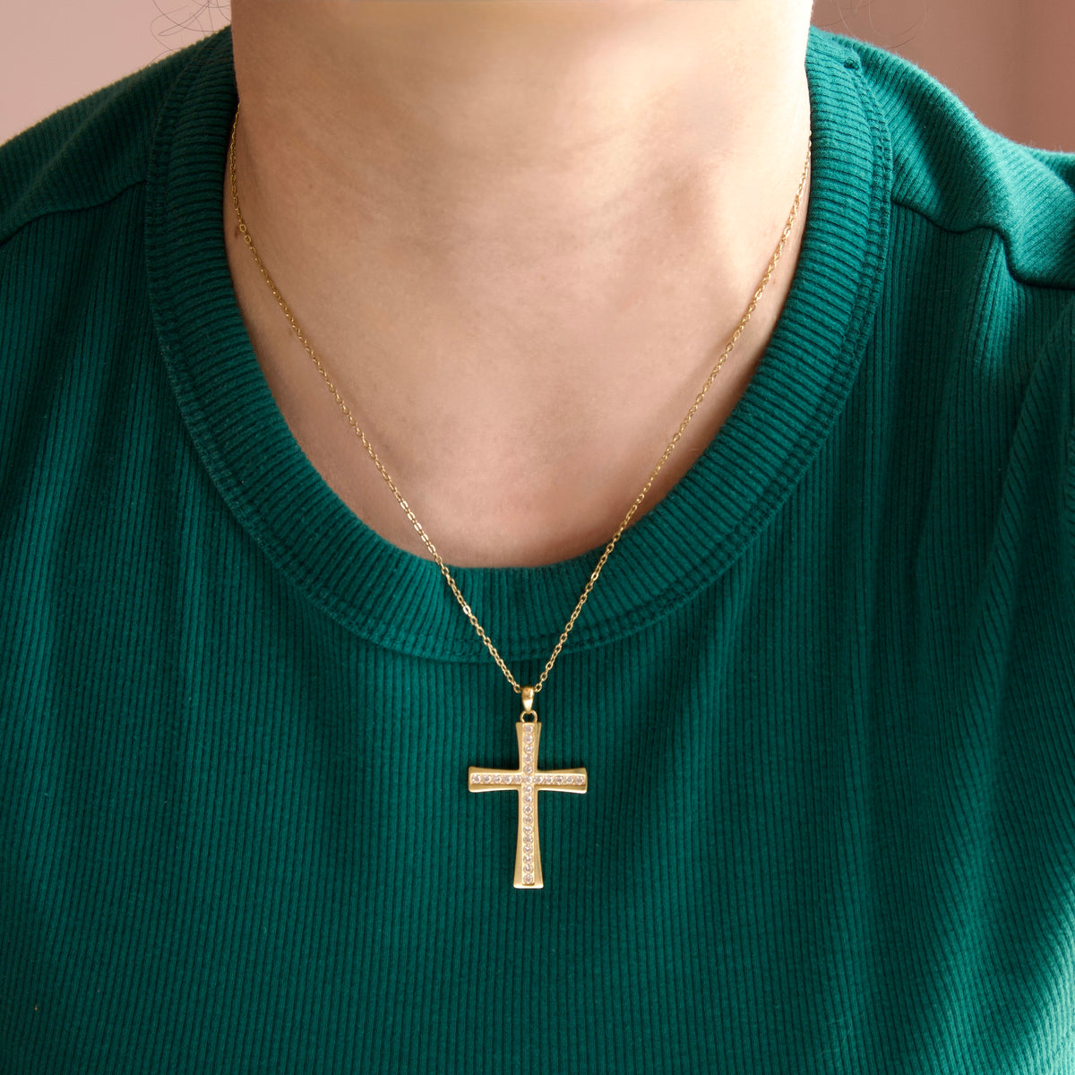 A gold cross pendant necklace with a textured, pave-like surface, worn by a person in a green ribbed shirt.
