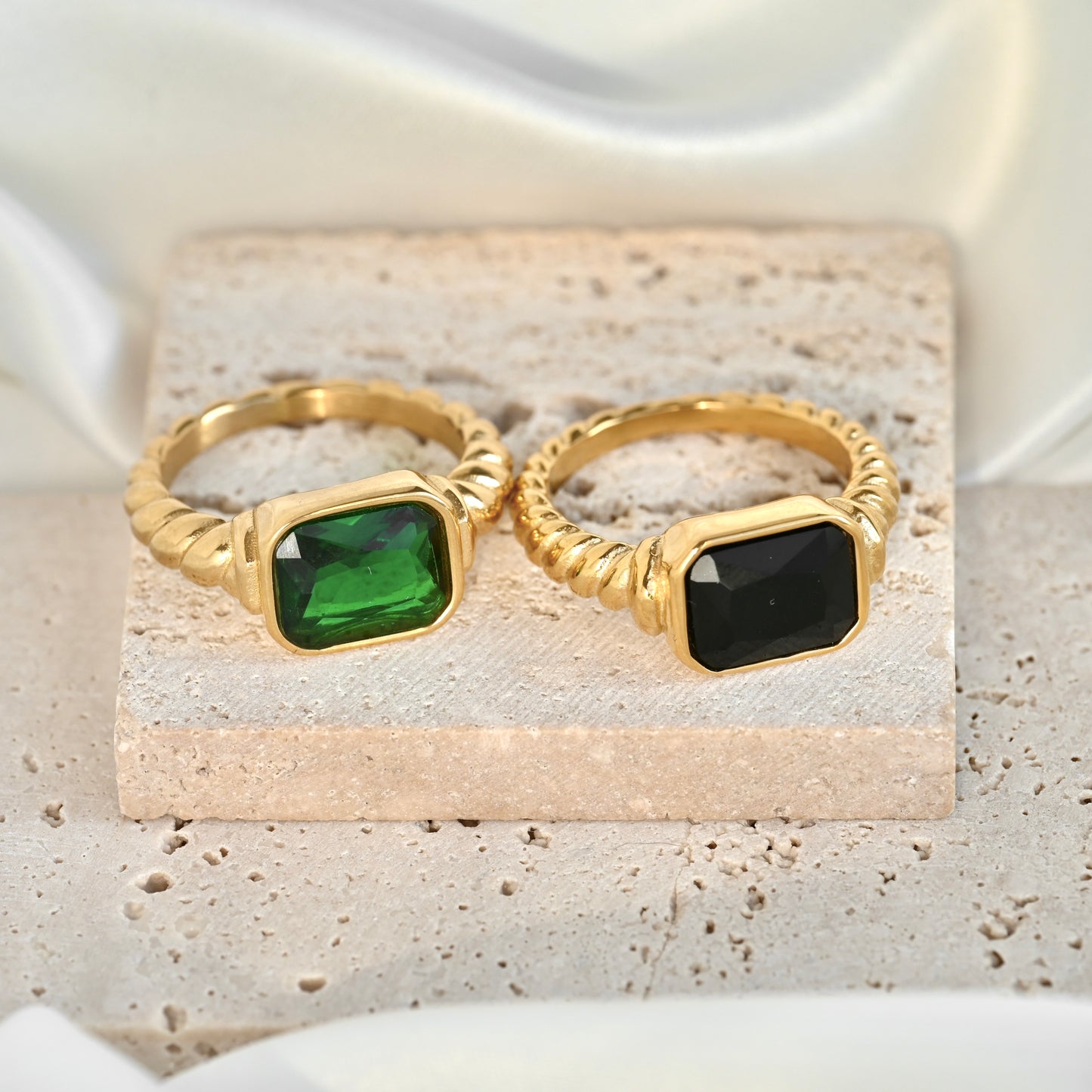 Two gold rings with rectangular green and black stones, displayed on a textured stone block with white satin fabric in the background.