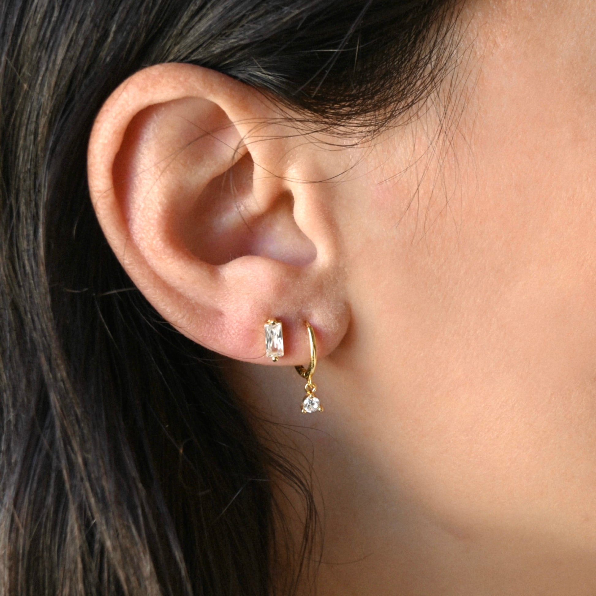 Close-up of an ear wearing gold hoop earrings  with CZ dangle in the first hole and baguette diamond studs in the second hole 