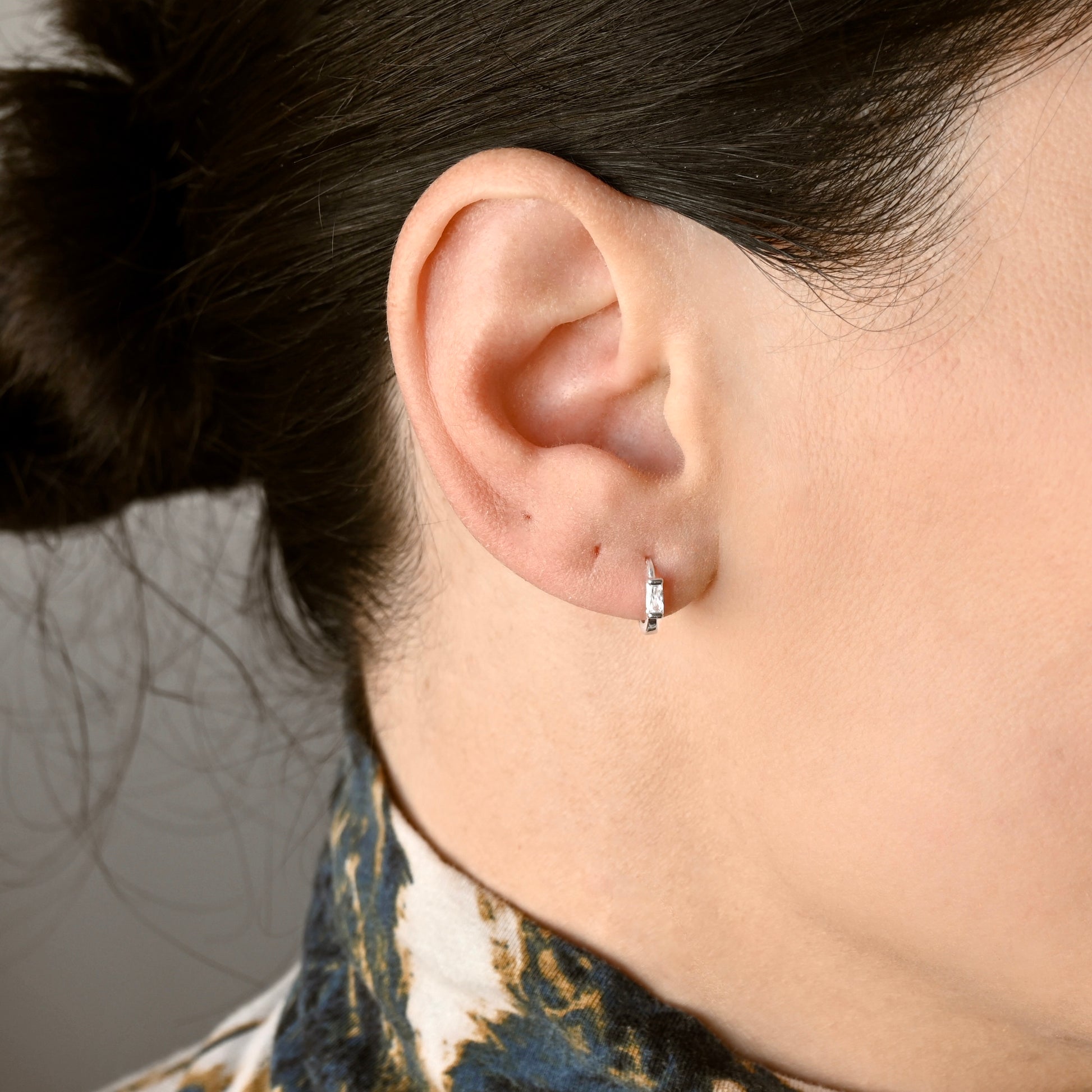 A close-up of a person's ear wearing a small silver hoop earring with a clear rectangular stone, against a neutral gray background.