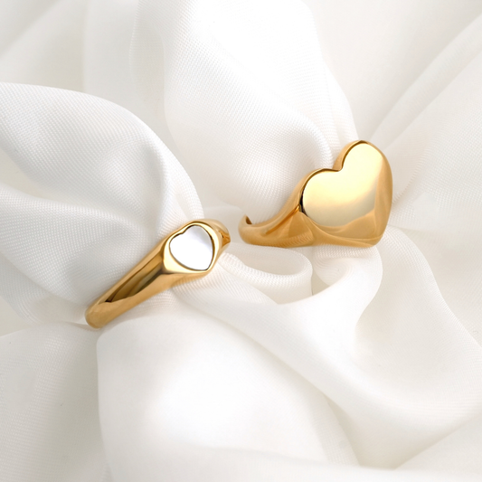 Two gold heart-shaped rings, one with shell inlay and the other being oversized signet style, on a white fabric background