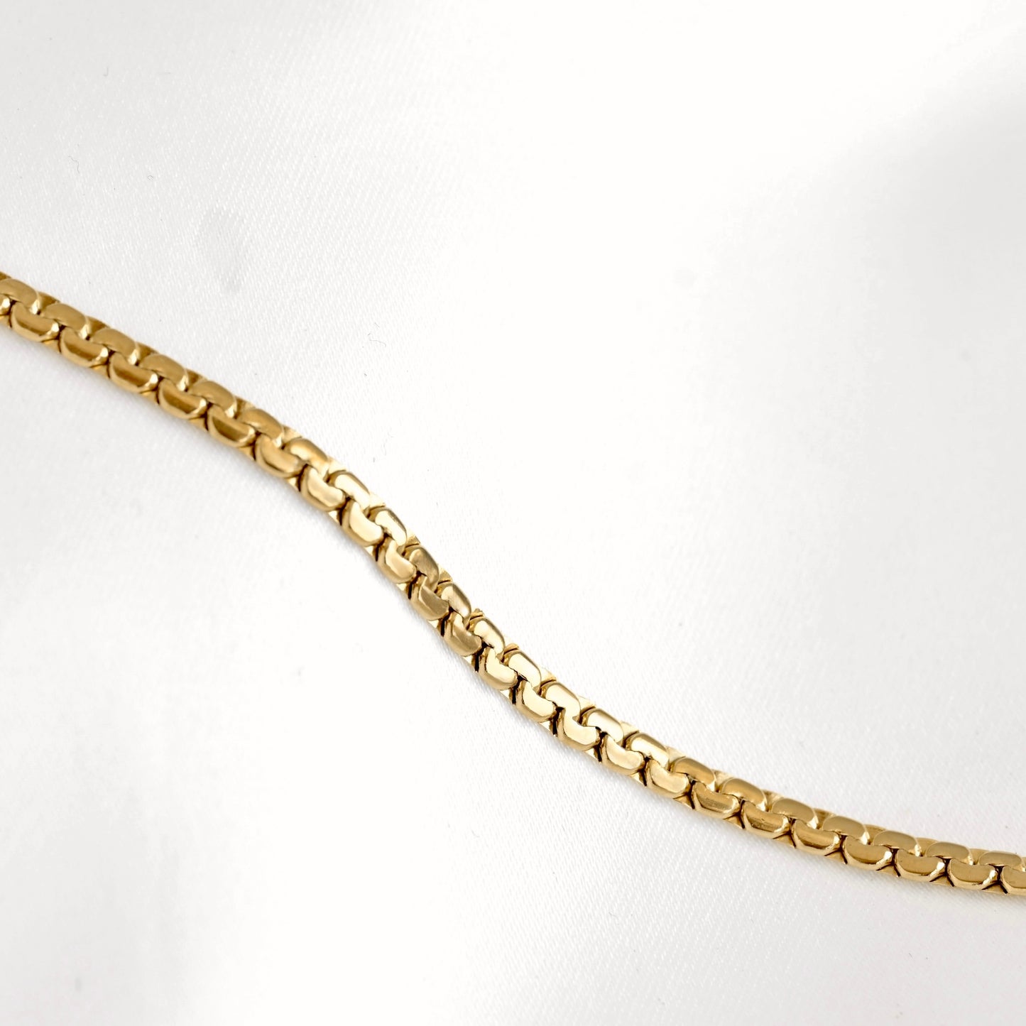 A close-up of a gold chain necklace with a flat curb link design, laid diagonally across a white textured background.