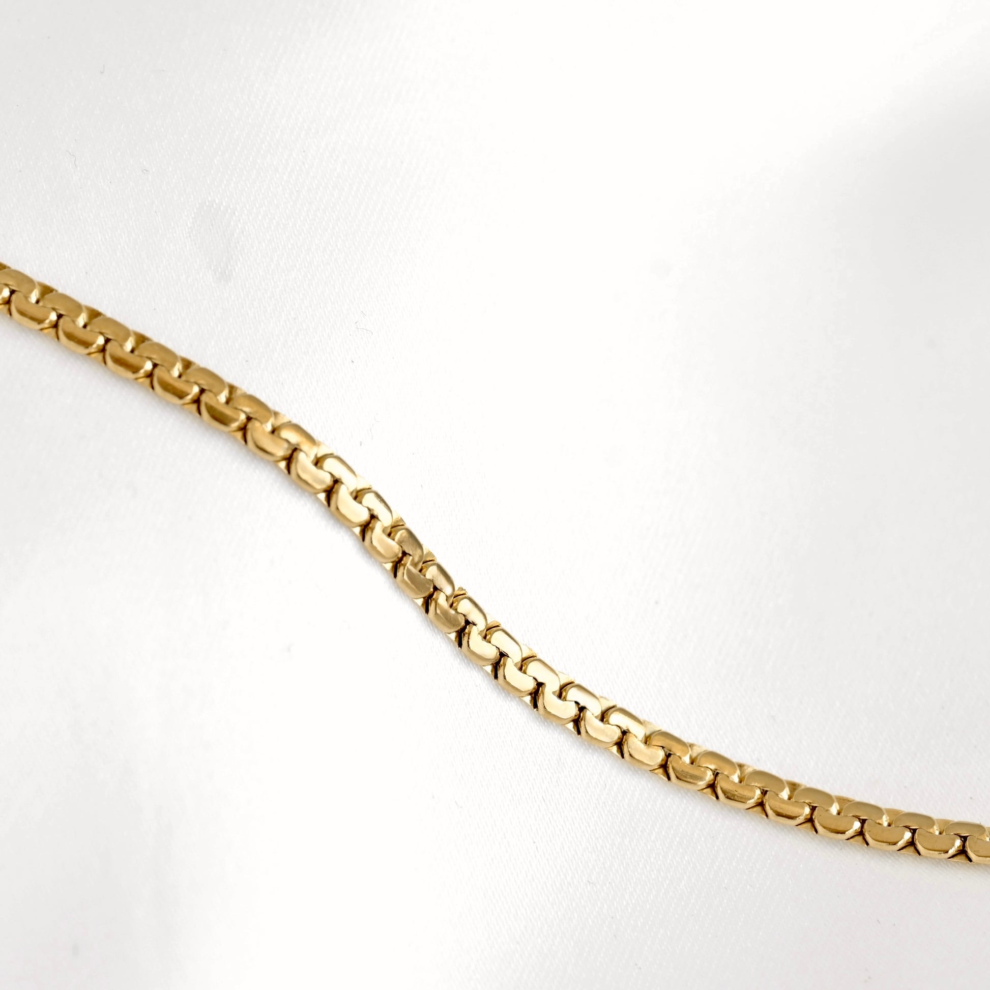 A close-up of a gold chain necklace with a flat curb link design, laid diagonally across a white textured background.