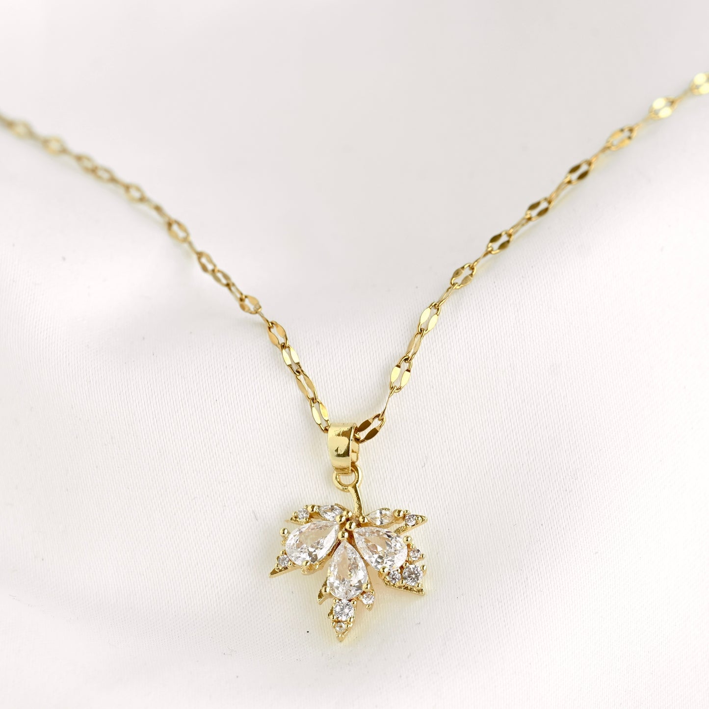 A gold chain necklace with a small leaf-shaped pendant set with clear stones, displayed on a soft white background.